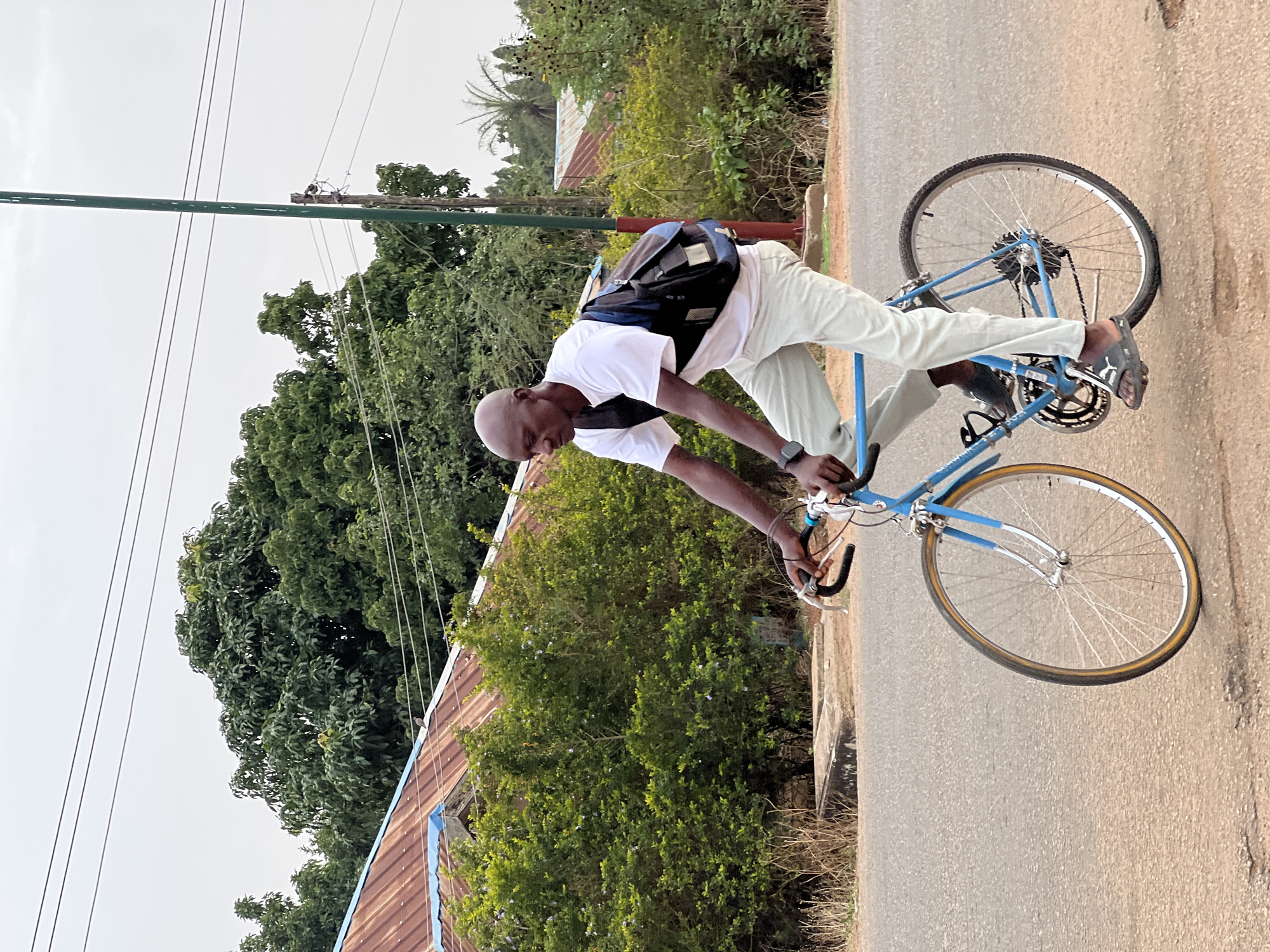A man is on a blue bicycle on a paved road in Bukuru, Nigeria. The man is bald, wearing a white shirt, light-colored pants, and a backpack. He is leaning forward, holding the handlebars. The bike is blue with silver wheels. The road is paved and cracked. Lush green vegetation borders the road. A building with a rusty roof is visible behind the vegetation. An electrical pole with lines extends into the sky. The sky is overcast.