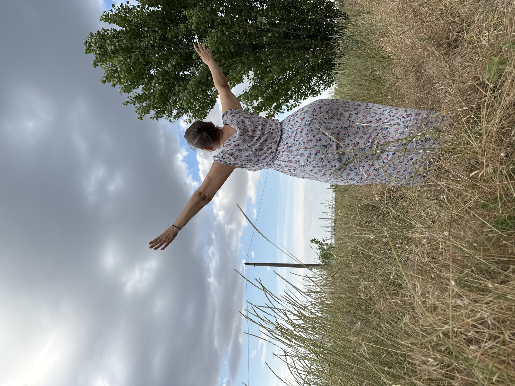 A person in a floral dress with their back to the camera stands in a field of dry grass, arms outstretched towards a cloudy sky and a large tree. The image is taken from a low angle, with the person's arms and the sky dominating the upper portion of the frame. A telephone pole is visible in the middle ground. The weather appears to be overcast with some patches of blue sky. The scene evokes a sense of freedom and connection with nature. The location is Odesa, Ukraine.