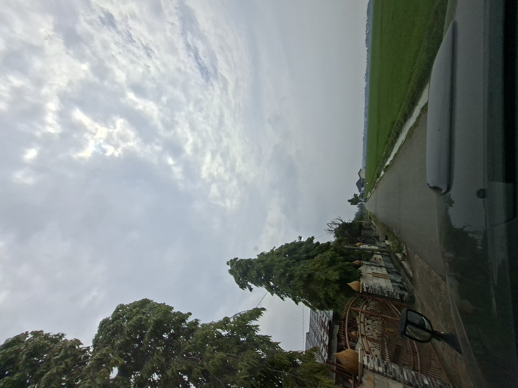 The image captures a view from a vehicle driving down a rural road, likely in Blangawe, Indonesia, under a cloudy sky. On the left side of the road, a decorative metal gate and a stone wall are visible, bordered by lush green trees. A tall, conical evergreen tree stands prominently near the gate. The road itself is paved and appears to be a two-lane street. To the right of the road, an expansive, vibrant green rice paddy stretches into the distance. A narrow irrigation canal runs alongside the paddy. In the background, several small houses and buildings are scattered among trees, and a distant treeline marks the horizon. The hood and side mirror of the vehicle are visible in the foreground, indicating the perspective of the photographer.