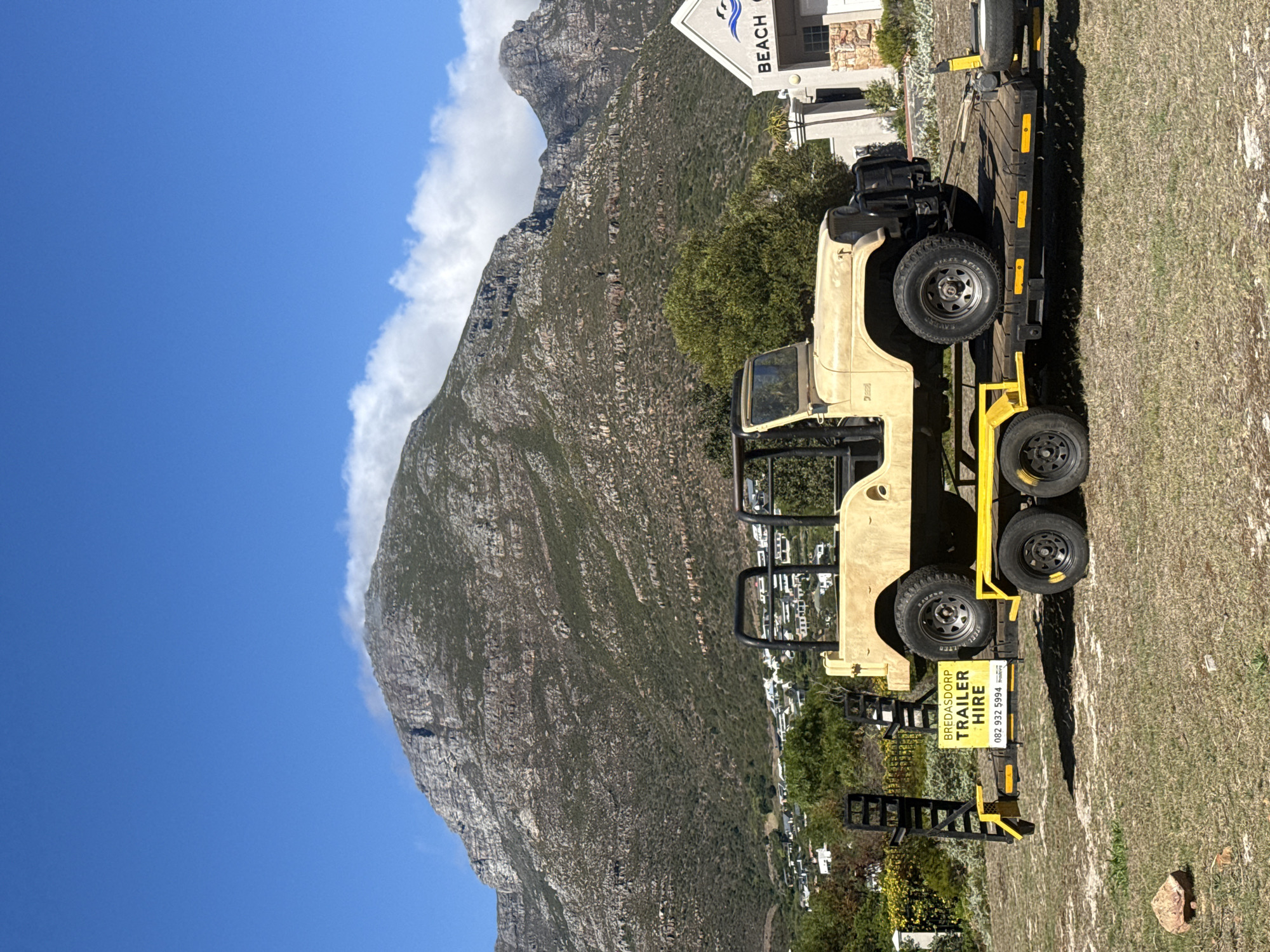 A beige Jeep-like vehicle without doors is secured on a yellow trailer, parked on a grassy area. A sign on the trailer advertises Bredasdorp Trailer Hire. In the background, a large mountain rises, partially obscured by clouds. Further behind the trailer is a building with a sign that says "BEACH". The scene appears to be in Hout Bay, South Africa, based on the visible landscape.
