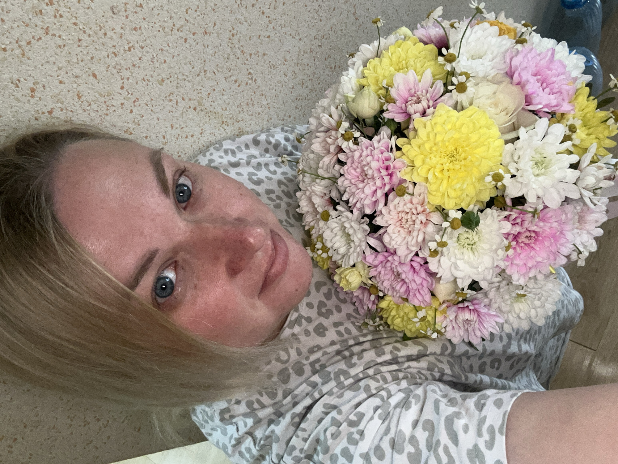 An overhead indoor shot captures a woman holding a large, vibrant bouquet of flowers. The woman, with blonde hair and striking blue eyes, looks up towards the camera. Her complexion is fair, with a slight flush on her cheeks and nose. She appears to be reclining or lying down, dressed in a light-colored garment with a distinctive grey leopard print pattern.
The bouquet, prominent in the foreground and held close to her, is a lush arrangement primarily composed of various types of chrysanthemums and possibly some smaller white daisies or chamomile. The dominant colors in the bouquet are bright yellow, crisp white, and an array of pink and light purple hues, creating a cheerful and full display.
The background consists of a light, textured surface, possibly a wall or floor, speckled with small brown or orange dots. In the upper right corner, a blurred blue object, which could be a water bottle or container, is partially visible. The lighting is soft and even, suggesting an indoor setting. There is no visible text in the image. The woman's expression is calm and content, hinting at a pleasant occasion, possibly receiving the flowers as a gift in Chaikovsky, Russia.