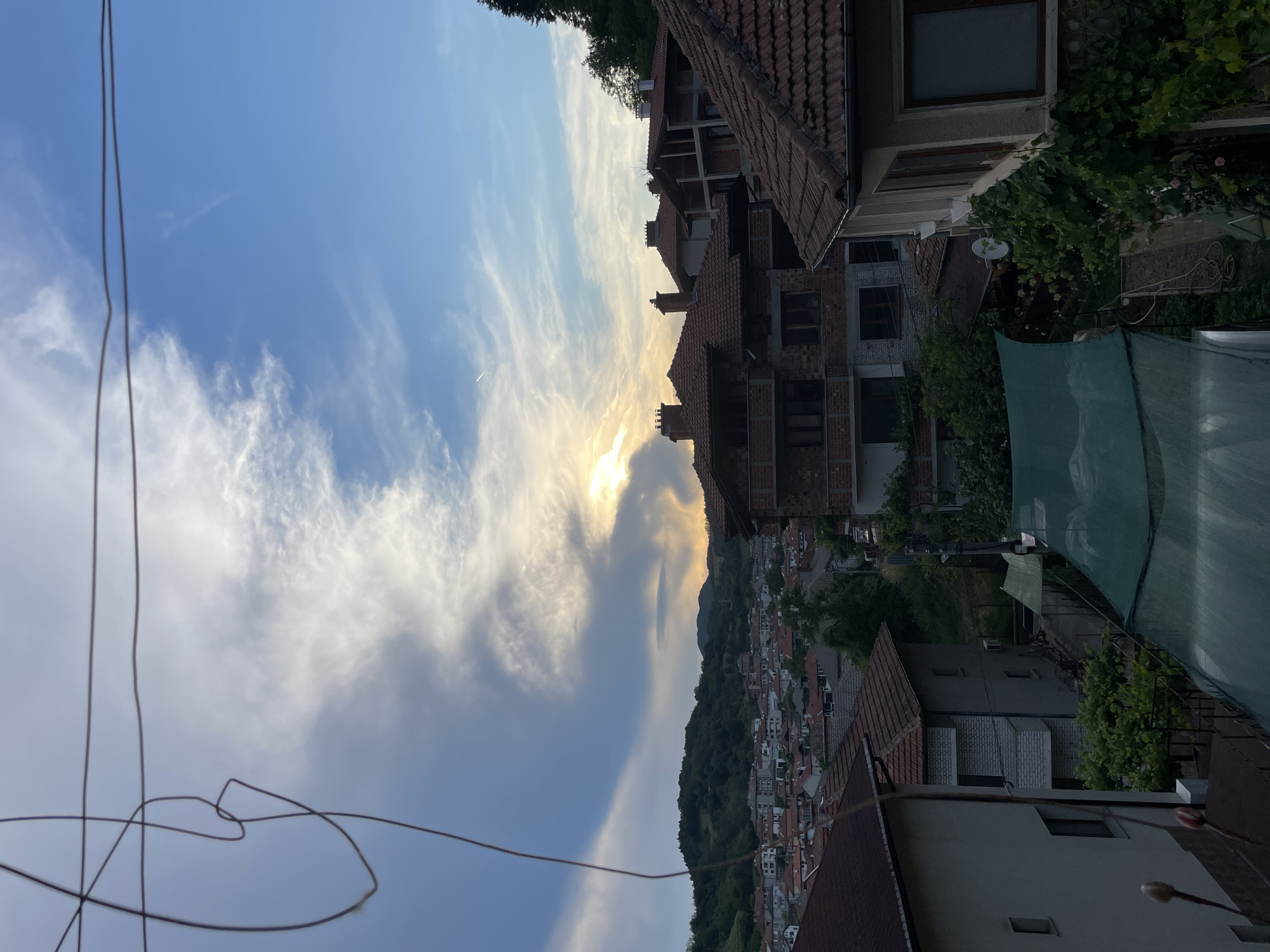 The media file is a vertical, low-angle outdoor photograph taken in Zlatograd, Bulgaria. The scene is dominated by a dramatic sky filled with wispy white clouds, with the sun breaking through the clouds and casting a warm, golden light. In the foreground, several taut wires stretch across the frame, creating a sense of depth and framing the scene.
Below the sky, a hillside town is visible, characterized by tightly packed houses with red-tiled roofs. The architecture suggests a traditional European village setting. The town extends down the slope of a lush, green hill, which is covered in dense foliage and trees.
The photograph appears to have been taken during the late afternoon or early evening, given the low angle of the sun and the warm, soft light. The weather seems pleasant, with clear skies and scattered clouds. There are no people visible in the frame. The overall mood of the image is serene and picturesque, capturing a quiet moment in a charming town. No text is visible in the media file.