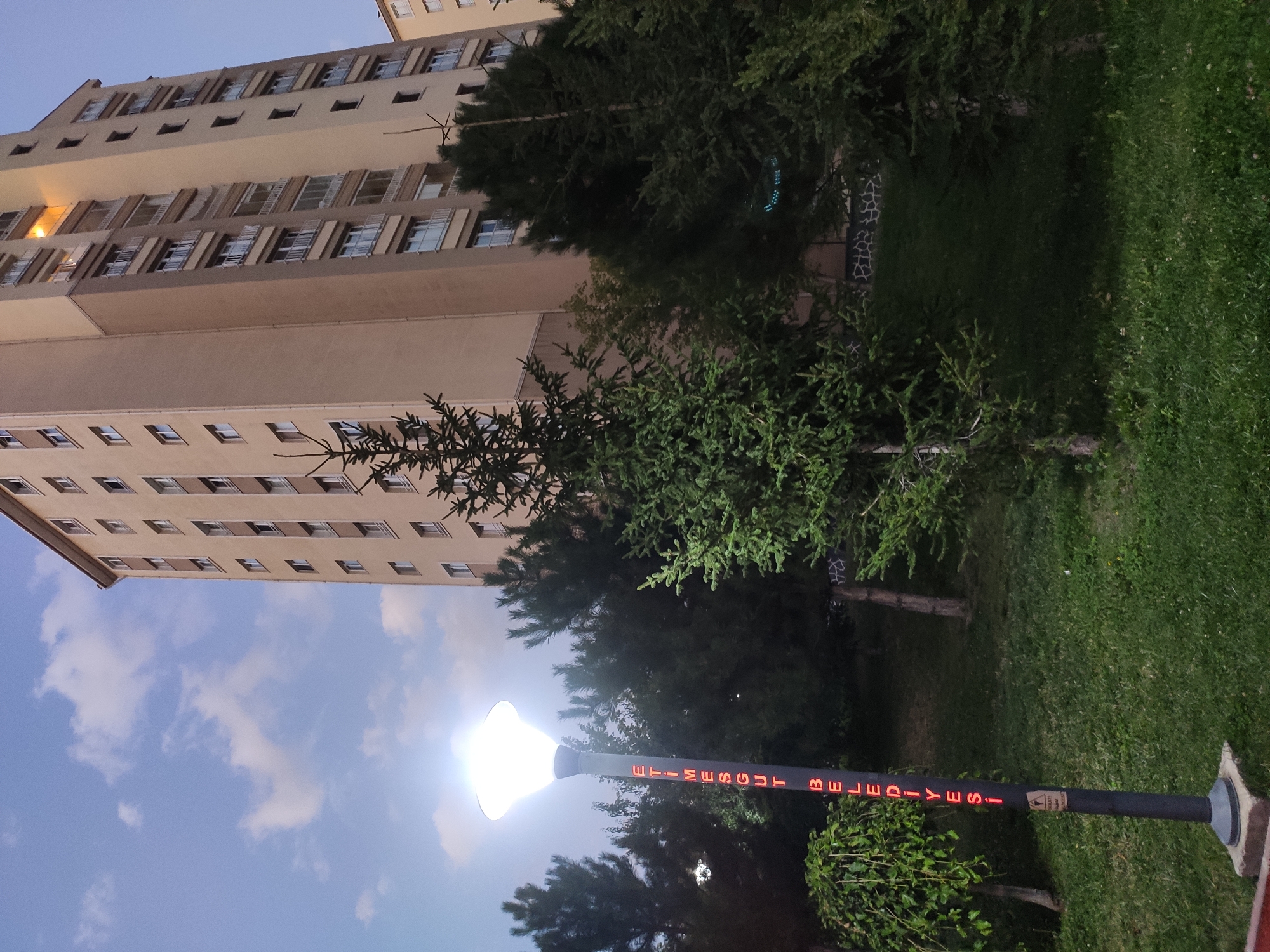 A tall, light-colored apartment building dominates the right side of the image, with multiple floors and windows visible. In the foreground, a lamppost with a bright light shines, and red text is illuminated vertically on its pole. The text reads "ETİMESGUT BELEDİYESİ", which translates to "Etimesgut Municipality". The lamppost stands amidst green grass and several trees, including some conifer trees that partially obscure the apartment building. The sky above is blue with scattered clouds. The scene appears to be an outdoor public space within the Etimesgut district of Ankara, Turkey.
