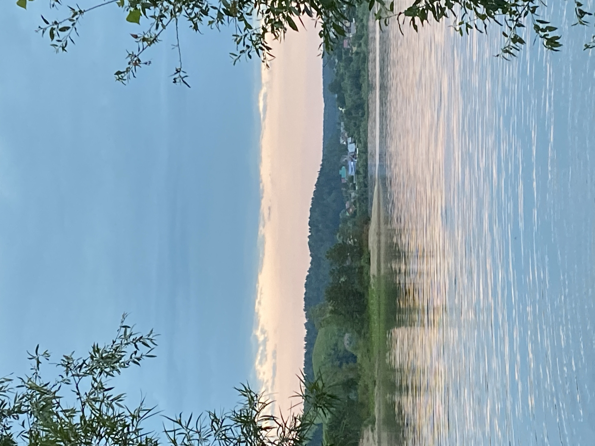 This is a horizontal, eye-level shot of a body of water, likely a river, in the city of Noviy, Russia. The water is calm with gentle ripples reflecting the sky and the distant shoreline. The foreground is dominated by the water’s surface, which transitions from a pale blue at the bottom to shades of pink and orange near the horizon, indicating either sunset or sunrise.
Framing the scene on the left and right are branches and leaves of trees, rendered in soft focus, creating a natural vignette effect. The leaves are a muted green, suggesting they are either backlit by the dawn or dusk light, or are of a species that has darker foliage.
The middle ground features a sandy bank leading to a lush, green shoreline. Beyond the shoreline, a rolling, wooded hill forms the backdrop. The hill is covered in dense, dark green trees. Nestled within the trees on the right side of the hill, a small settlement of houses with colorful roofs is visible.
The sky above is a gradient of pale blue, with subtle bands of muted pink and peach clouds low on the horizon, reinforcing the impression of either early morning or late evening. The overall mood of the image is serene and tranquil, capturing a quiet moment in a natural landscape with a touch of human habitation.