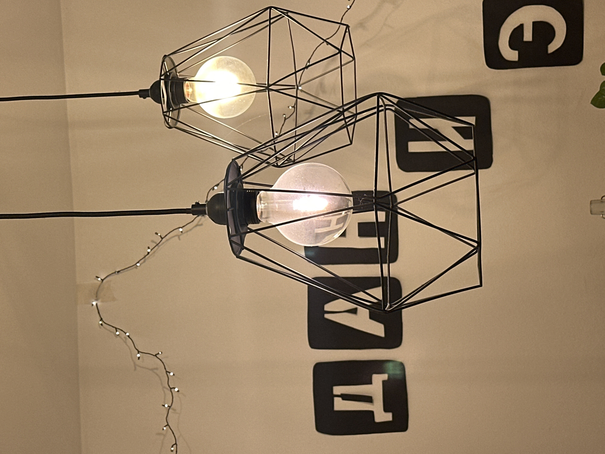 Two hanging light fixtures with geometric wireframe shades are illuminated against a light beige wall. The upper light fixture is slightly out of focus, while the lower one is sharper. To the right of the light fixtures are four black squares, each containing a white letter: "E," "A," "T," and "T." A string of small decorative lights runs down the left side of the frame. The location is likely indoors in Ramat Gan, Israel. There are no visible people, actions, or interactions in the image. The focus is on the inanimate objects and their arrangement.