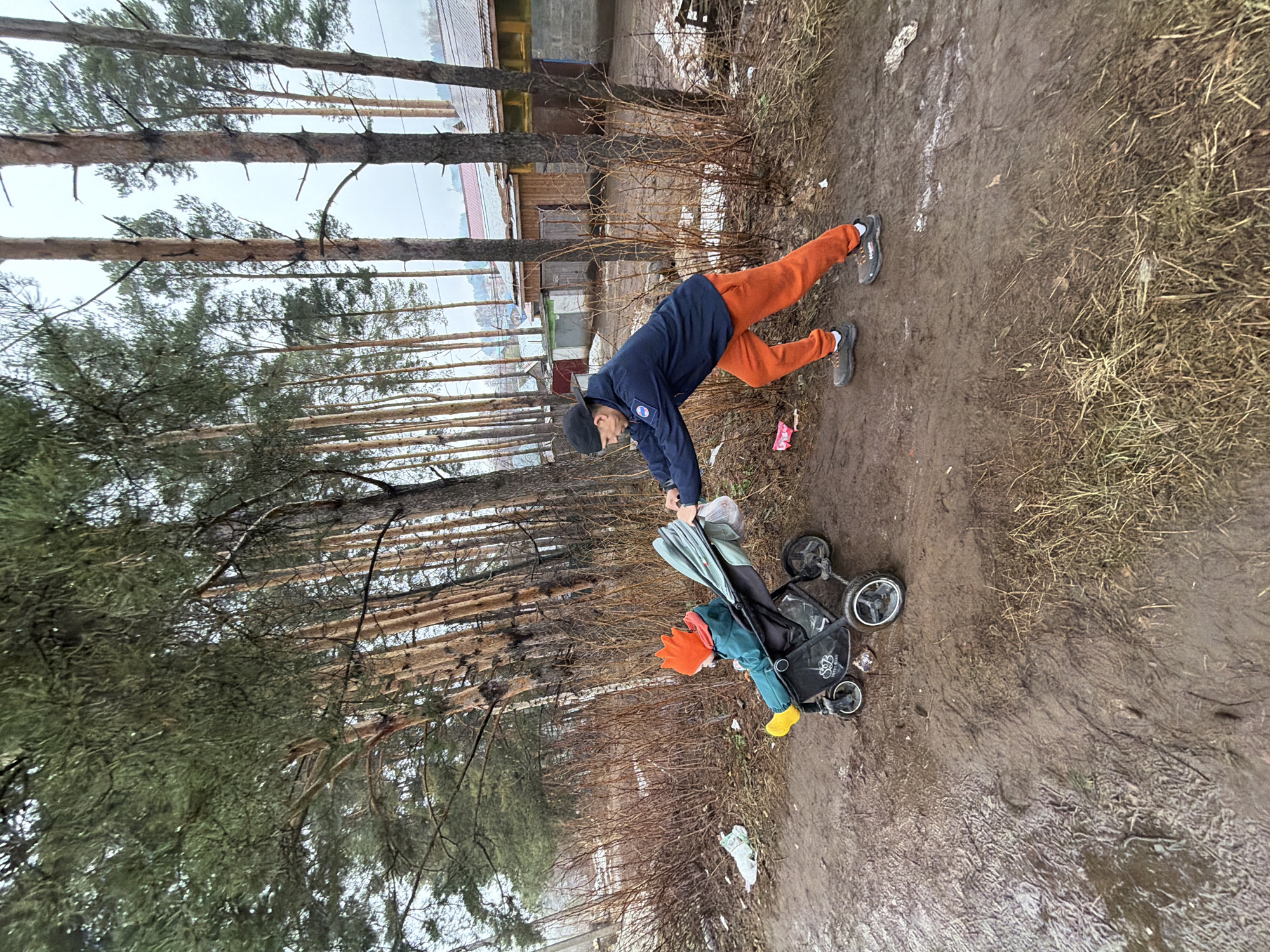 A person dressed in orange pants and a blue jacket, wearing a black cap and gray shoes, is pushing a stroller on a muddy path. The stroller contains a child dressed in green with an orange hat and yellow boots. The path is surrounded by tall, thin trees and some brush. A few small buildings are visible in the background. The location is Chaikovsky, Russia.