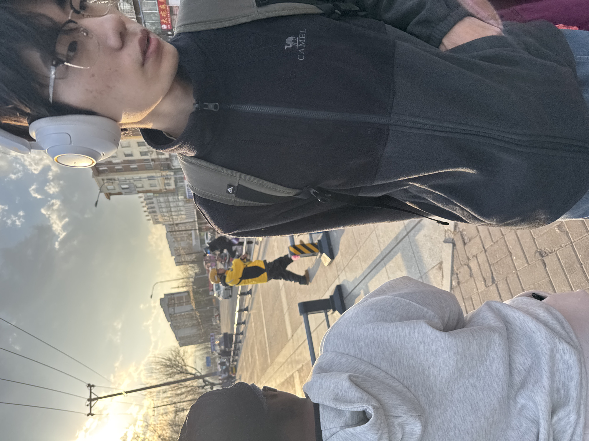 A street scene in Zhongtun, China, captured from a slightly canted perspective, features a young person wearing headphones and glasses, and a black "CAMEL" fleece jacket. In the background, a road construction worker is visible in a yellow and black uniform. Buildings line the street under a bright sky.