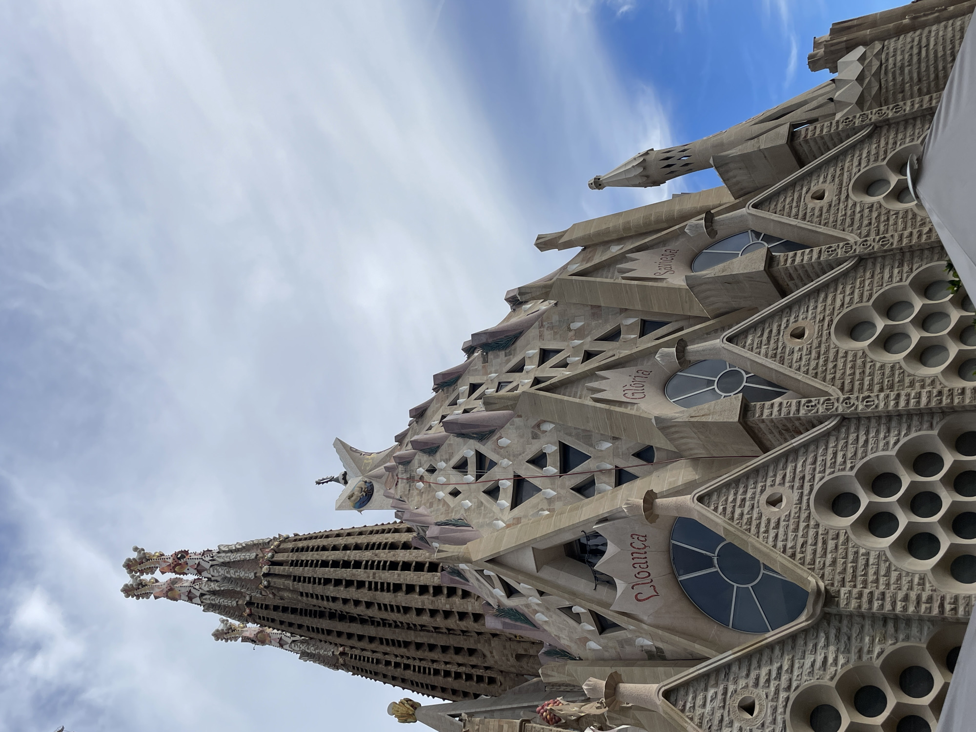 The image captures an upward view of the Sagrada Familia basilica in Barcelona, Spain. The structure's architectural details are prominent, including the honeycomb-like textures, triangular windows, and the words "Gloria" and "Lloança" inscribed. A tower rises in the foreground, showing its intricate construction. The sky is partially visible, with a mix of blue patches and scattered clouds.