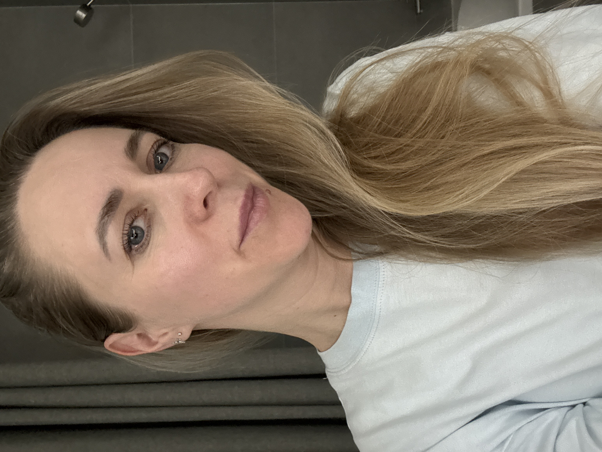The image is a portrait of a woman with long, flowing blonde hair and blue eyes, captured in an indoor setting. She has fair skin and a light complexion, displaying a natural, neutral, and slightly pensive expression as she looks directly at the camera, likely in a selfie. Her hair is spread out, resting on a surface behind her, suggesting she might be lying down or leaning back.
She is wearing a light-colored, possibly light blue or white, long-sleeved crew-neck top. A small, decorative ear cuff or hoop earring is visible in her left ear. The background primarily consists of dark, possibly grey, tiled walls or panels, providing a muted backdrop. In the upper left portion of the image, a metallic fixture, resembling a shower head or a minimalist ceiling light, can be seen. To the right of her head, a white, soft object, likely a pillow or bedding, is visible. The scene is set indoors, in Chaikovsky, Russia. There is no discernible text present in the image.