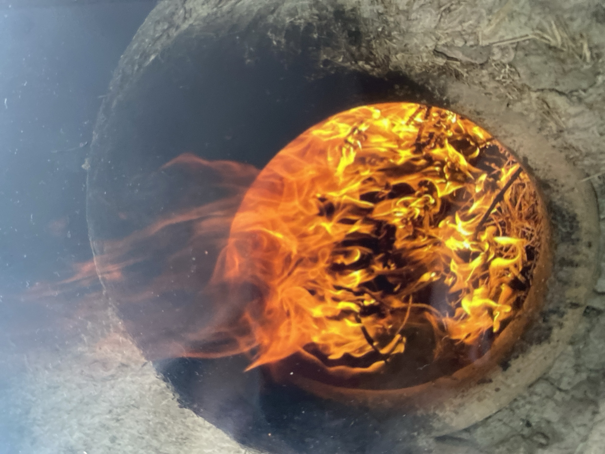 An overhead view shows a circular opening in a rough, greyish-brown material, possibly earth or clay, with a prominent, bright orange and yellow fire burning within the opening. The fire is composed of licking flames and appears to be consuming dry, straw-like material. The inside of the opening, where the fire is contained, is darker and appears to be the interior of a pit or oven structure. The outer material is textured and uneven, with some lighter colored debris scattered around.