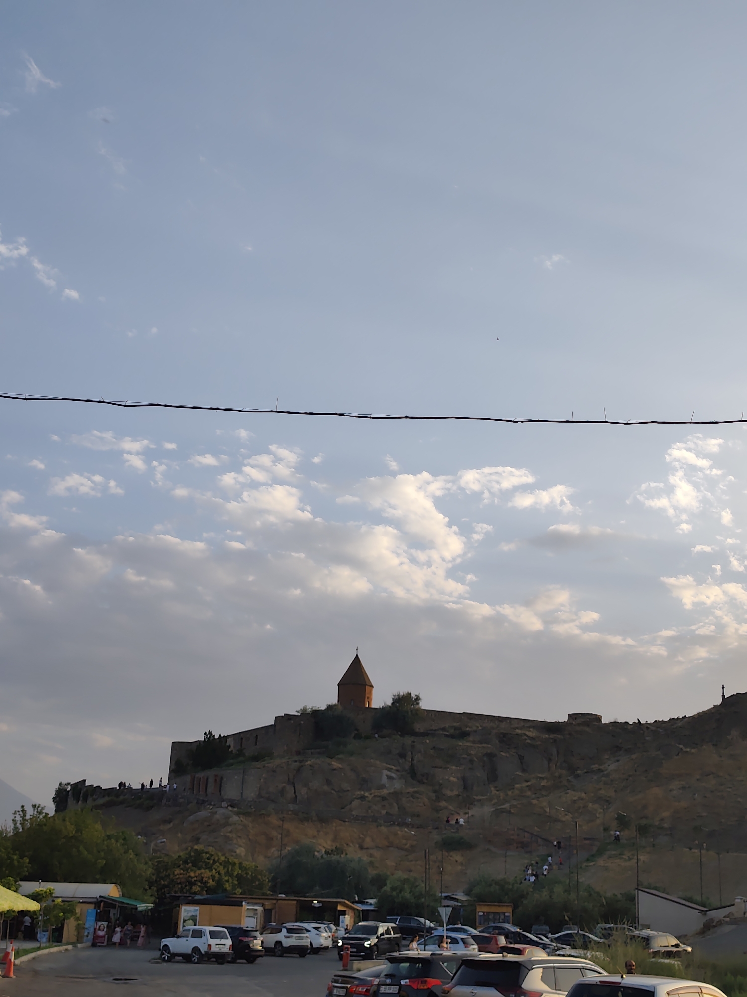 The image depicts a view of the Khor Virap monastery in Lusarat, Armenia. The monastery is situated atop a rocky hill, with its distinctive conical-roofed church visible. A wall surrounds the monastery complex, and several figures are seen walking along the ramparts. Below the hill, a parking lot is filled with various cars. In the foreground, several small market stalls with yellow awnings are set up. The sky above is a pale blue with scattered white clouds, and a power line is strung across the upper portion of the frame. The overall scene appears to be from a late afternoon or early evening, given the soft lighting.