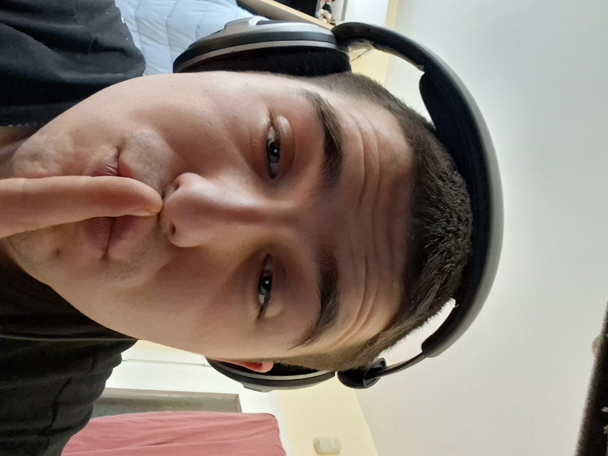 A close-up, angled portrait shows a man wearing headphones and holding a finger to his lips. He appears to be indoors, possibly in a residential setting, given the suggestion of a bed in the background. The location in the city of Plovdiv, Bulgaria is not determinable from the visual information provided.