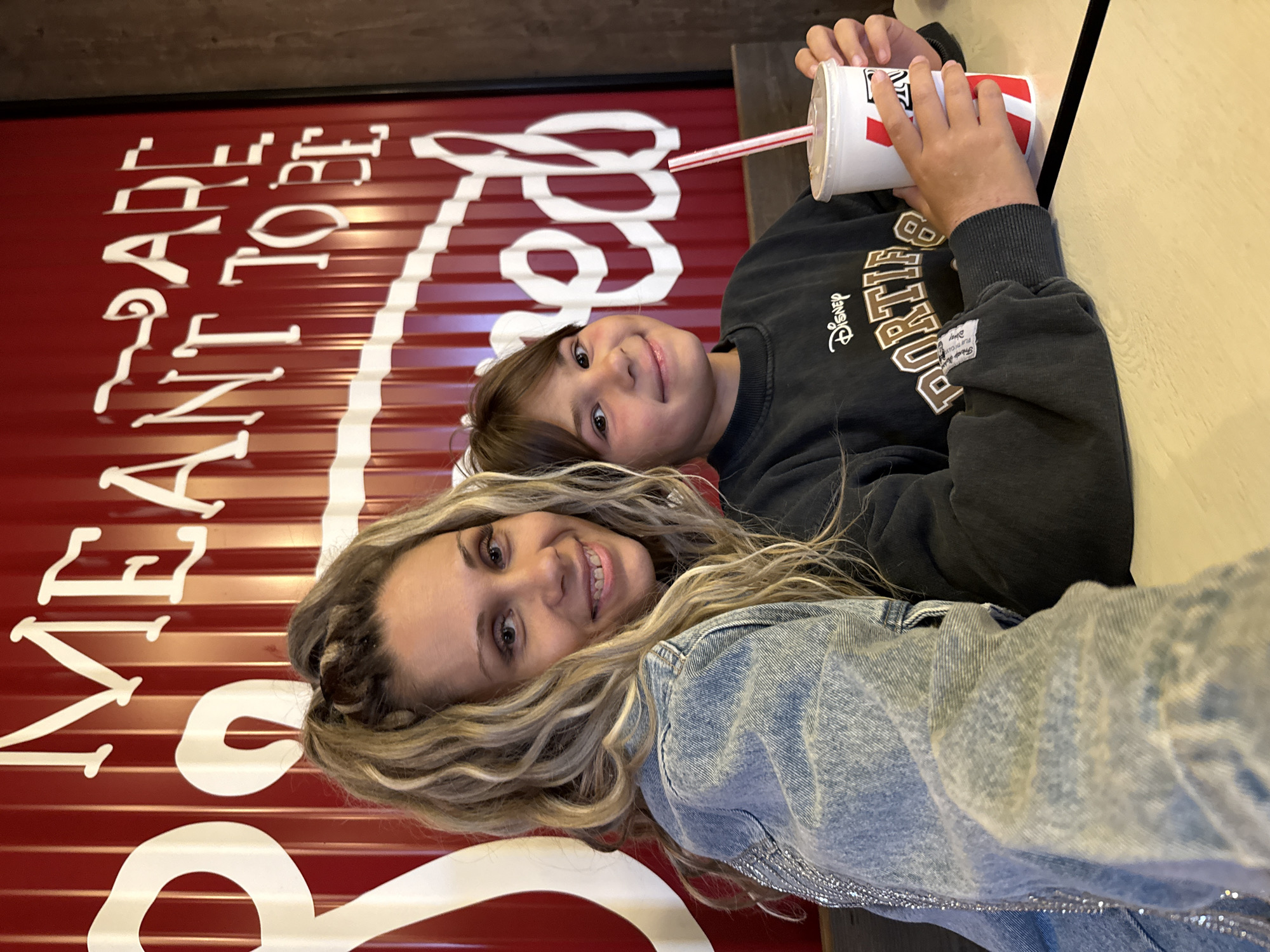 A medium shot shows a woman and a young boy sitting at a table with a red, corrugated metal wall in the background that has white lettering on it. The woman is on the left and is wearing a denim jacket. Her hair is blonde and styled in a high ponytail. She is smiling at the camera. The boy is on the right and is wearing a dark sweatshirt. He is holding a white cup with a red straw in it. He is also smiling at the camera. The table appears to be made of light-colored wood. The overall lighting suggests an indoor setting. This image was taken in Ignatovo, Russia.