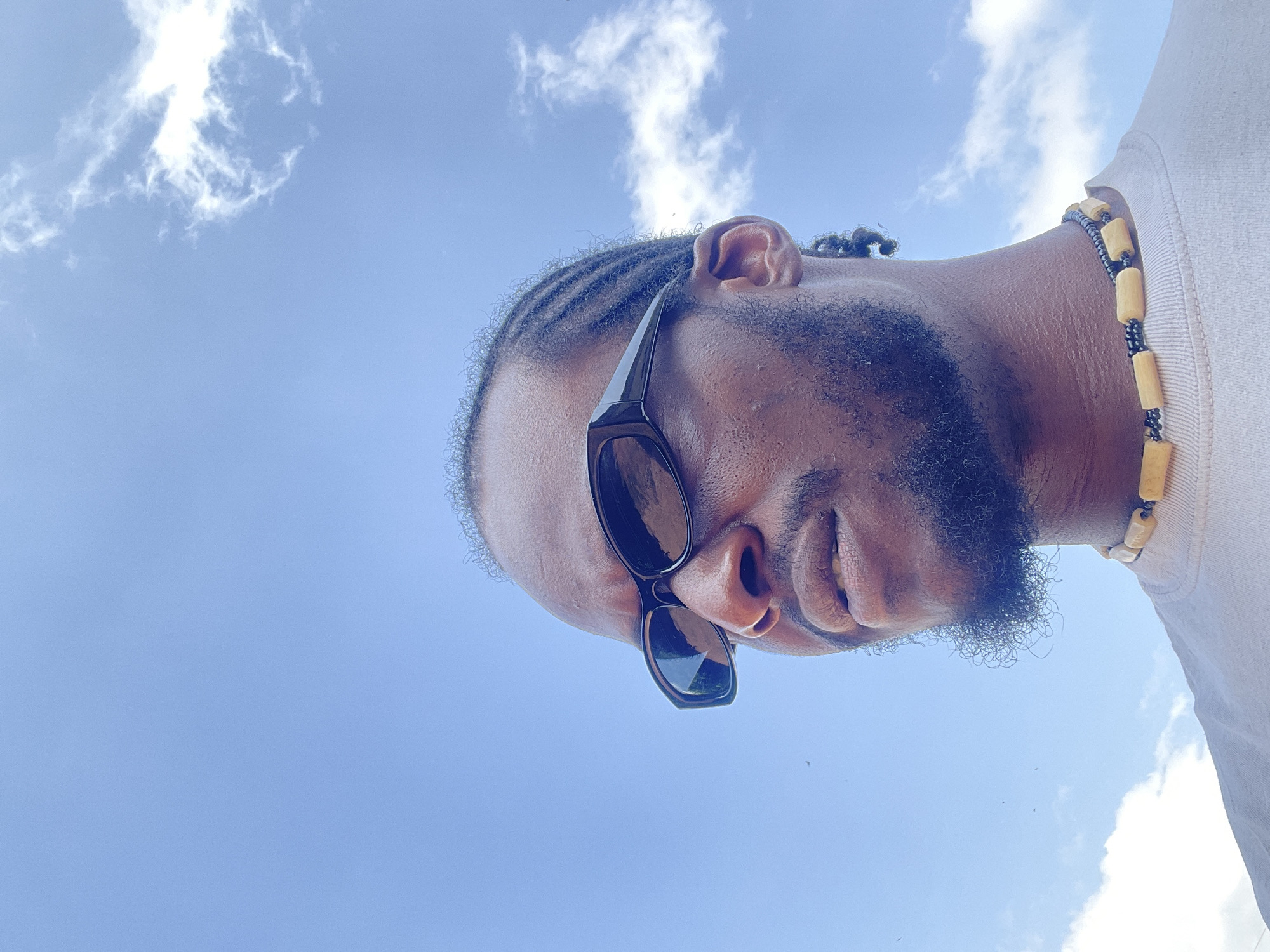 The image is a low-angle, close-up shot of a man from the chest up, taken outdoors against a bright blue sky with scattered white clouds. The man is wearing black rectangular sunglasses and a light-colored t-shirt. He has braids in his hair, a beard, and is wearing a beaded necklace made of light-colored, cylindrical beads and black beads. His gaze is directed to the left of the frame. The scene appears to be in an open, outdoor environment, consistent with a location in the city of Jos, Nigeria. The lighting suggests it is daytime.