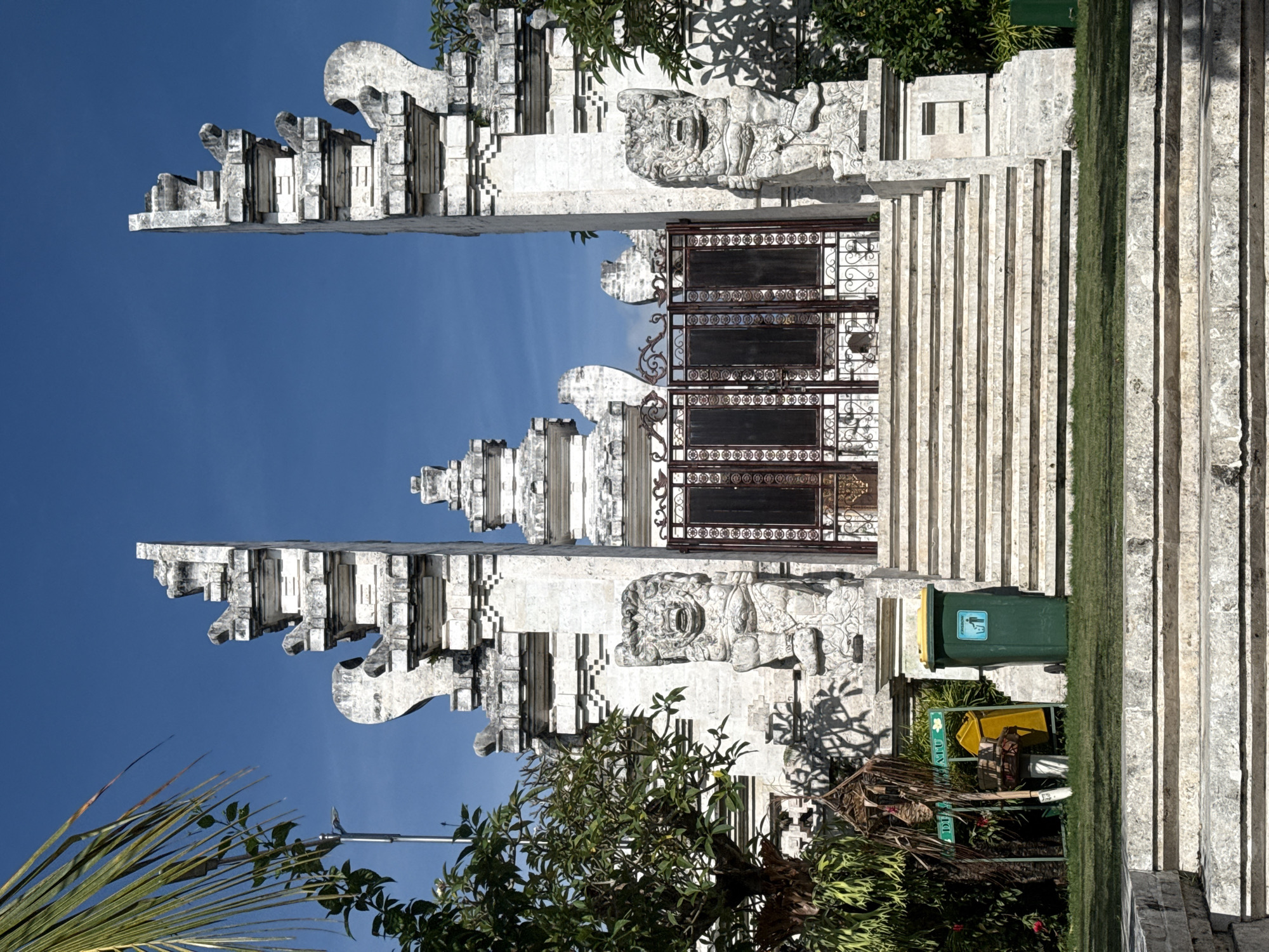 The image depicts a traditional Balinese gate, a Candi Bentar, in Pecatu, Indonesia. The gate is constructed of light-colored stone or concrete and features intricate carvings and a split design, leaving an open passage in the center. A wrought iron gate is visible behind the candi bentar. To the right of the gate, a series of stone steps leads upwards, bordered by a strip of green vegetation. A green waste bin with yellow lid and a yellow container can be seen near the steps. The sky is clear and blue. A palm frond is visible at the lower left. The image is rotated ninety degrees counter-clockwise.