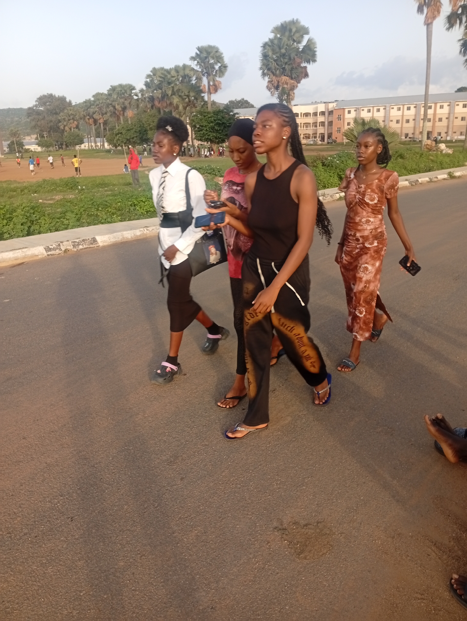 The image depicts a group of four young women walking on an asphalt road in Yelwa, Nigeria. The women are walking in the same direction, from left to right, and appear to be engaged in conversation or simply walking together.
The woman in the front on the right is wearing a sleeveless black top and black pants with white lettering. She has long, dark braided hair. Next to her, slightly behind and to her left, is a woman in a patterned brown dress. She is also holding a dark-colored mobile phone in her right hand. To the left of these two, a woman in a white collared shirt and black skirt is visible. She has her hair pulled back and appears to be looking down at something in her hand. The fourth woman, mostly obscured from view, is in the foreground on the far left.
The environment suggests a semi-urban or suburban setting. In the background, there is a field with people playing what appears to be soccer. Beyond the field, there are rows of palm trees and buildings that look like residential or educational structures. The sky is clear and appears to be late afternoon or early evening, given the lighting and the long shadows. The road has a white painted line on its edge.