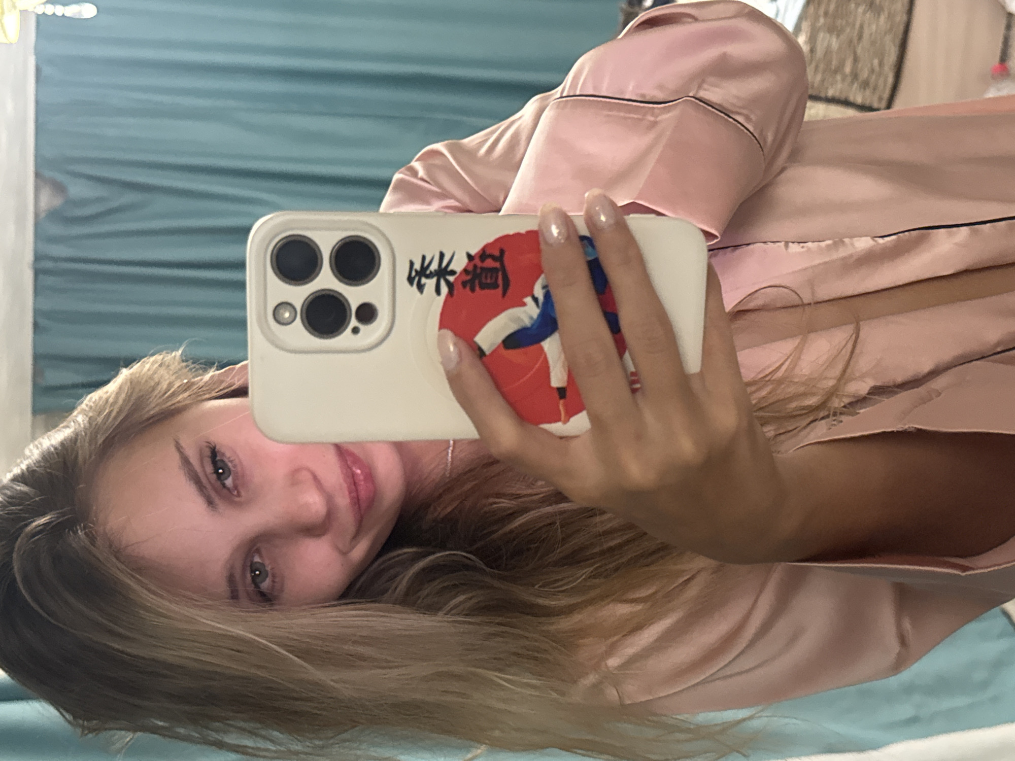A selfie-style image depicts a woman with light brown hair and fair skin, lying on a light blue surface (possibly a bed or couch). She is wearing a pink satin pajama-style top. She is holding a white smartphone with a distinctive camera array (likely an iPhone) and a sticker featuring a red circle with what appears to be a person in a white gi and black Japanese characters. The background includes a light blue curtain. There is no information to determine the location of the picture in Chaikovsky, Russia.
