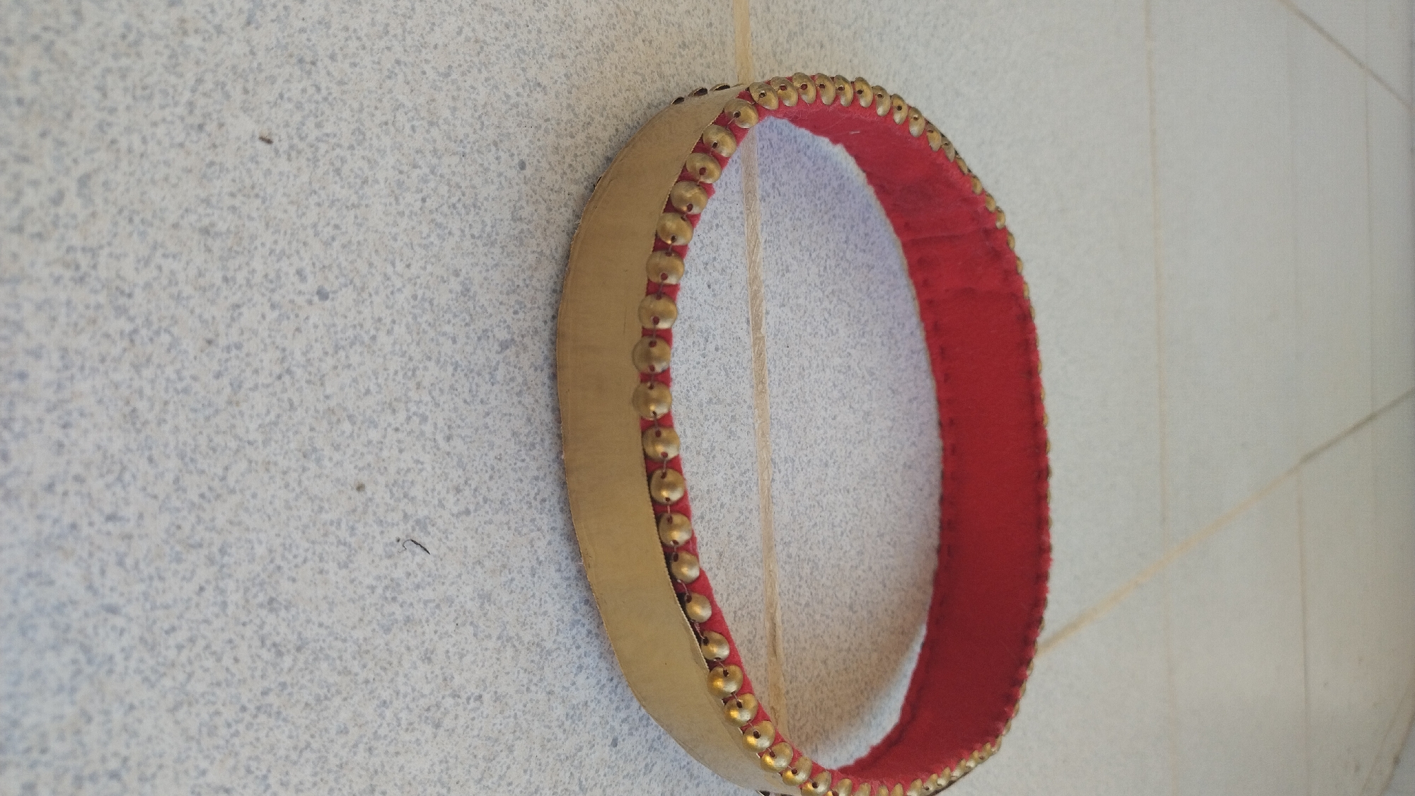 A close-up, eye-level shot shows a decorative gold and red circular object placed on a light gray tiled floor. The object is a circular band with a red inner lining and a wider gold outer band. The gold outer band is embellished with a continuous row of small, brass-colored studs along its upper edge, which faces the viewer. The red inner lining is visible along the inner circumference of the object. The tiled floor has a speckled pattern of light gray and darker gray tones. The lighting suggests an indoor environment, possibly in Perea, Greece, though no specific urban landmarks are visible. No people or actions are depicted.