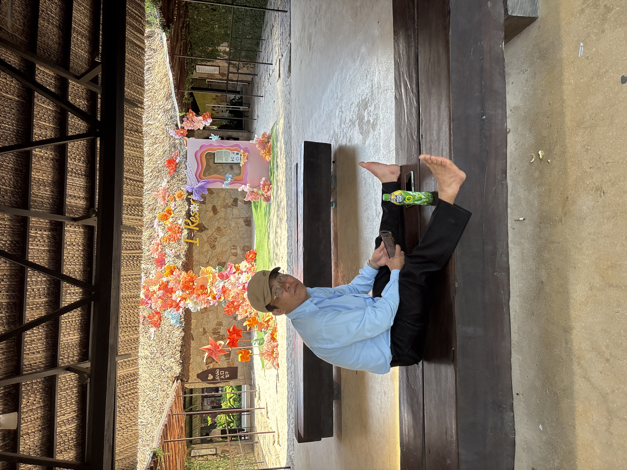 A man is sitting on a wooden bench, outdoors, under a thatched roof. He is wearing a light blue button-down shirt, black pants, a light brown baseball cap, and glasses. He is holding a smartphone in his right hand and a green bottle with a yellow label is on the bench to his left. His feet are bare, with his left foot resting on the bench and his right foot extended. Behind him, a wall is decorated with a large floral arrangement of red, orange, and pink flowers. A pink photo frame cutout with the words "#happyha... resortnhanhtrang" is visible in the center of the floral display. The ground is paved with concrete and sand. The location is identified as Tay Nha Trang Ward, Vietnam.