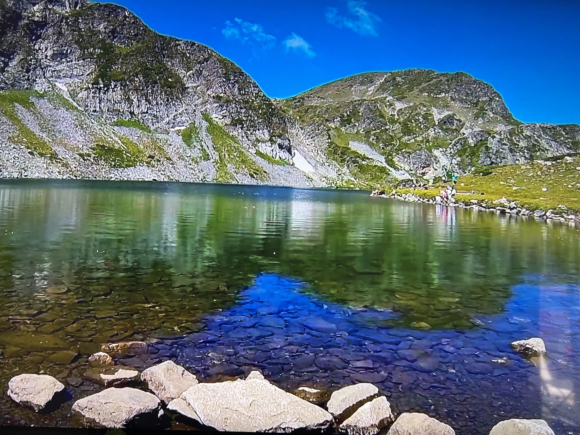The image depicts a mountain lake on a clear day. The foreground features several light-colored rocks partially submerged in the clear water. The water's surface reflects the blue sky and the surrounding mountains, creating a blend of colors. In the middle ground, a lake stretches towards the back of the image, with mountains lining its edge. Patches of green vegetation dot the mountain slopes. A small group of people are visible on the lake's edge on the right side of the frame. The sky is a bright blue with a few scattered white clouds. The image appears to be a photograph or a still from a video of a natural landscape. The location is not apparent from the image.