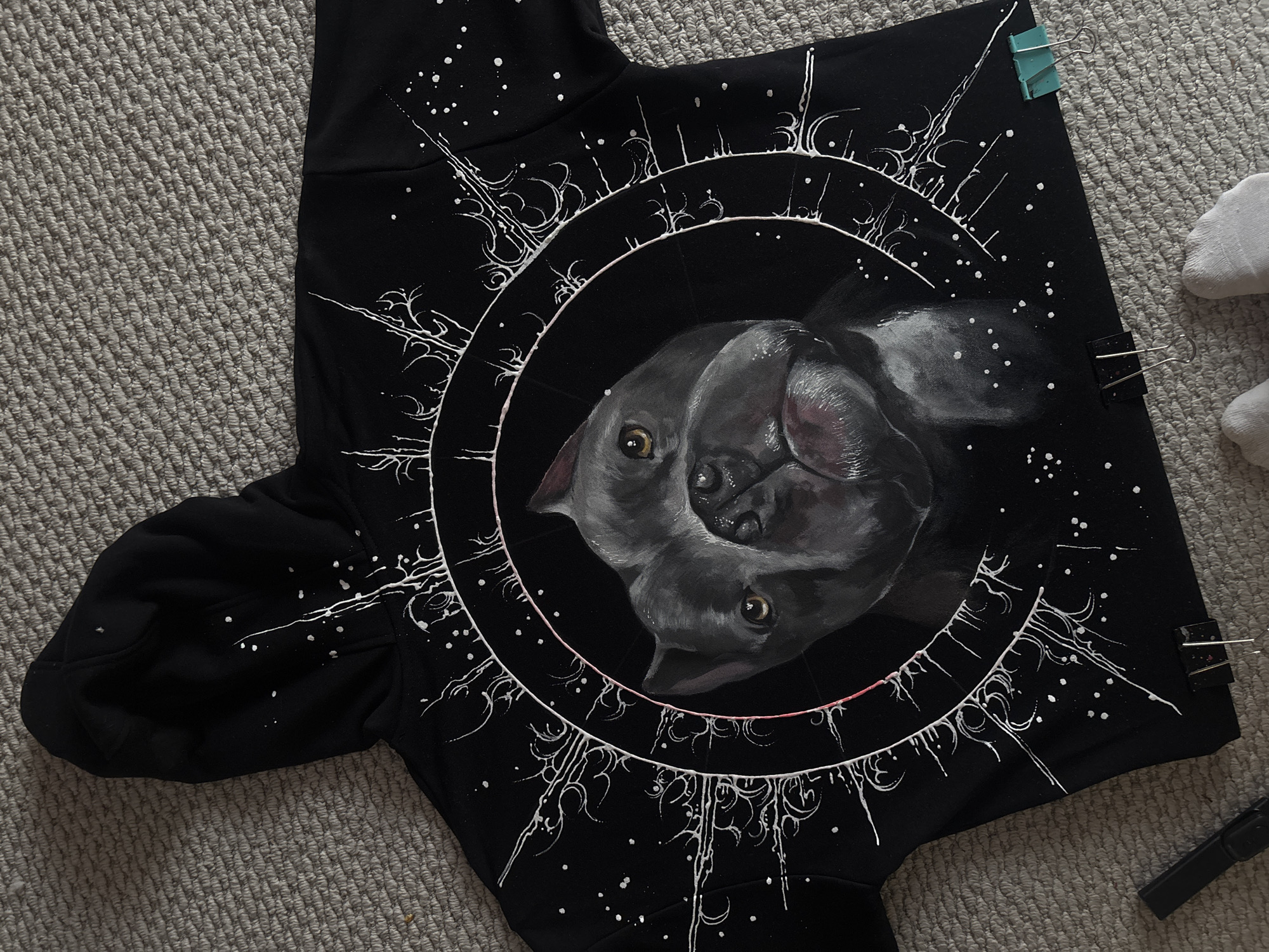 A black hoodie, adorned with a detailed hand-painted design, lies flat on a light grey, textured carpet. The central feature of the design on the hoodie's back is a realistic portrait of a grey or black dog's head, possibly a French Bulldog, with prominent brown eyes, depicted within a circular frame. The dog's expression is attentive, with its head slightly tilted.
Surrounding the central dog portrait are intricate white patterns resembling abstract roots, branches, or cosmic formations, interspersed with numerous white dots that suggest stars or a starry sky. These patterns radiate outwards from concentric circles around the dog's head. A thin, faint pink line is also visible tracing part of the inner circle. The painting appears to be done with thick, slightly textured paint.
The hoodie is positioned with its hood at the top left and sleeves extending to the sides. Two binder clips, one light blue and one black, are used to secure the top right edge of the fabric to an unseen surface or simply hold it flat. To the far right, parts of white socks or slippers are partially visible, resting on the carpet. A black pen or marker lies near the bottom right corner of the hoodie.
The image captures an indoor setting, likely a home in Krasnoyarsk, Russia, where a fabric art project has been recently completed or is in progress, with the painted hoodie laid out, possibly for drying or display. No specific time of day or emotional cues are directly depicted, and there is no visible text within the image.