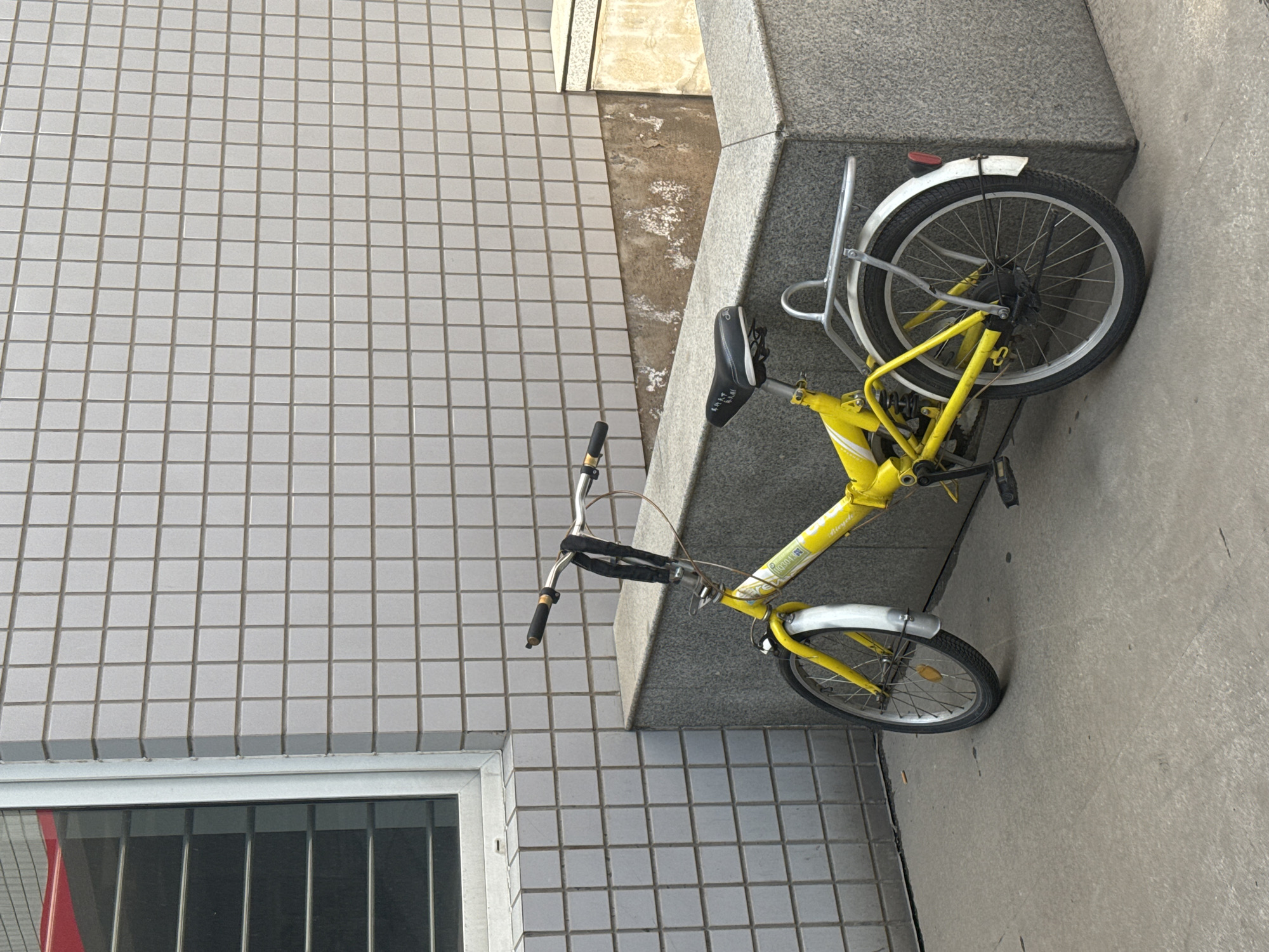 A yellow bicycle is parked on a sidewalk next to a granite retaining wall and a building with tiled walls in Zhongtun, China. The bicycle is angled slightly, with the front wheel closer to the building and the rear wheel nearer to the sidewalk edge. The seat of the bicycle is black. A window is visible on the lower-left side of the building. The sidewalk is concrete.