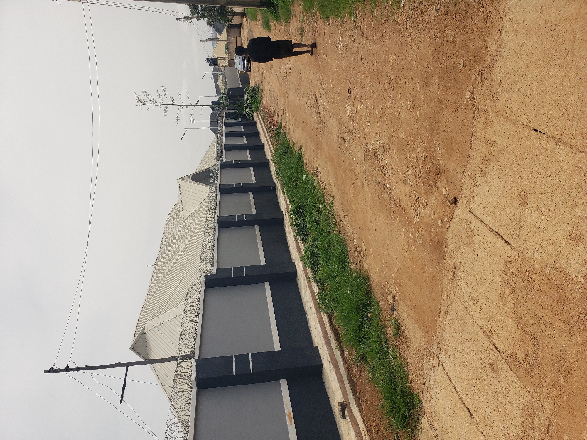 A single person, dressed in black, walks away from the camera down a dirt road in Jos, Nigeria. The person is positioned in the middle of the road, facing away and slightly to the right. To the left of the road is a tall, dark, and gray fence topped with barbed wire. The fence encloses a property with a silver-colored roof. A utility pole with wires stands to the left of the fence. Across the road and further down, a dark-colored car is visible. The sky is overcast, and the environment appears to be a residential area or a road leading to one. The ground is uneven, with patches of grass and some concrete slabs visible on the left side of the road.