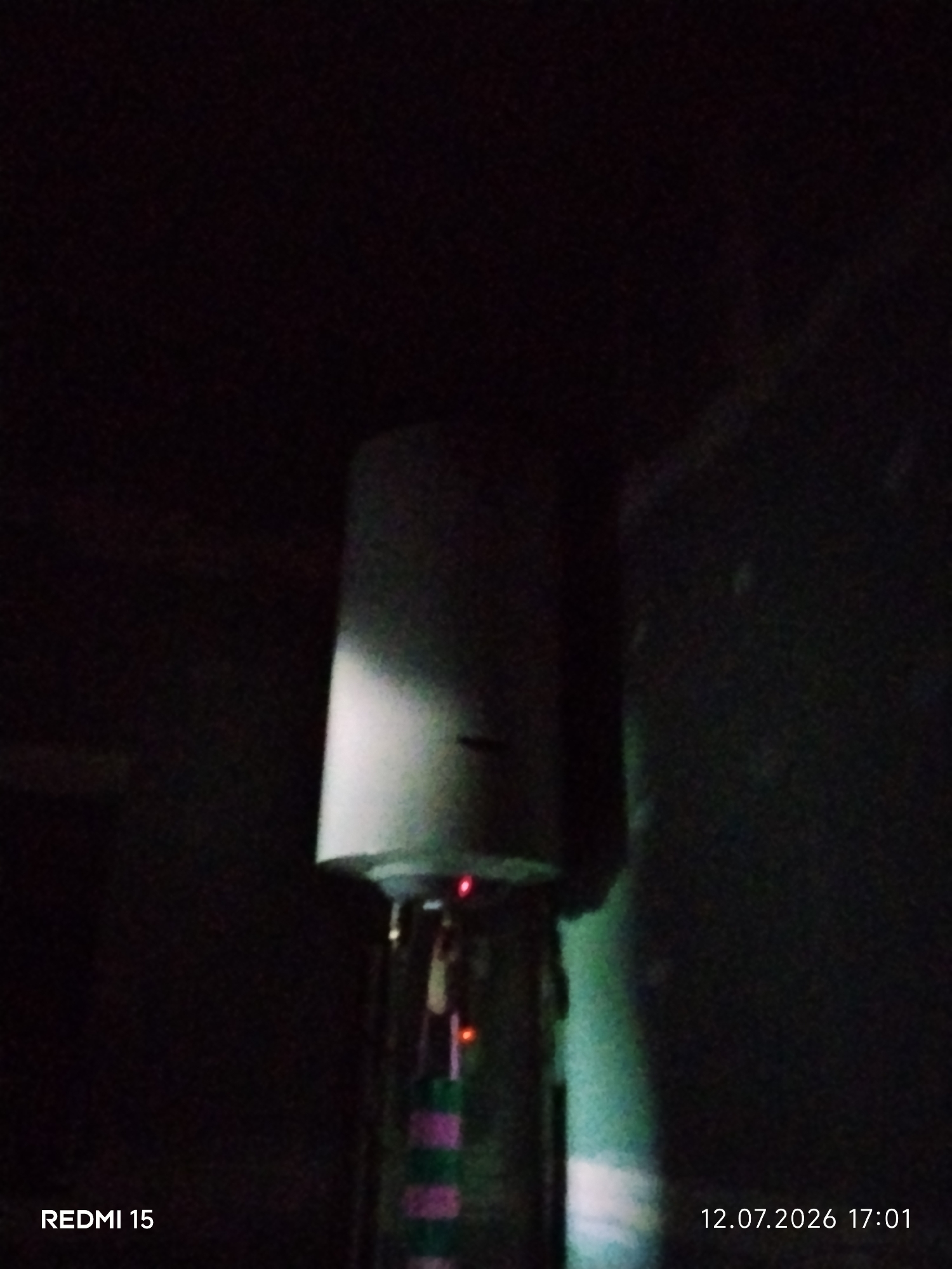 The image displays a vertical, cylindrical object, possibly a water heater or a piece of industrial equipment, set against a dark, underexposed background. A small red light is visible near the top of the object. The bottom portion of the object has horizontal stripes of pink and green. Text overlays in the bottom left corner read "REDMI 15", and in the bottom right corner, "12.07.2026 17:01". The lighting suggests an indoor or dimly lit environment. There is no clear indication of a specific location within the city of Ayiet, Kazakhstan.