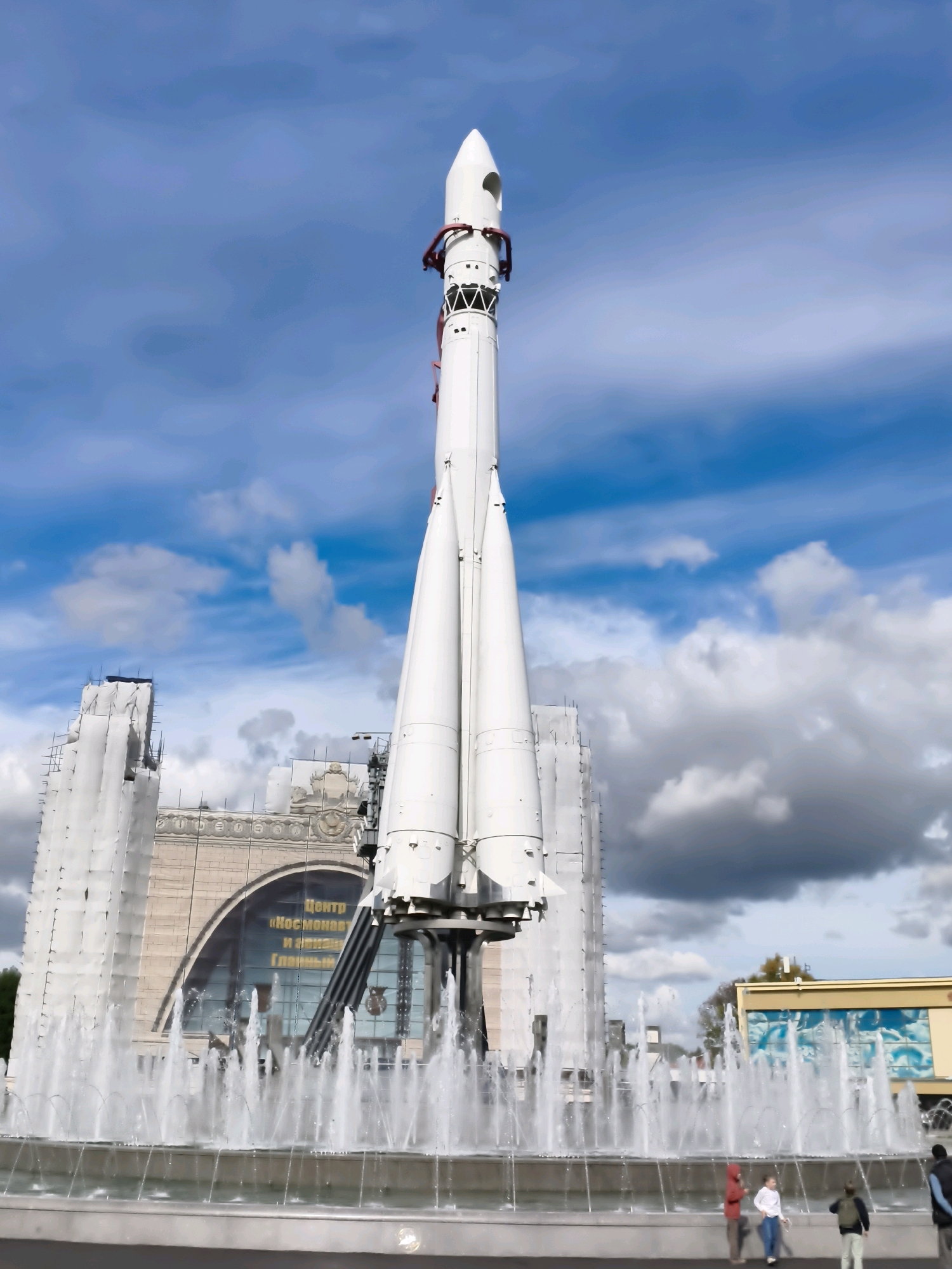 The image shows a full-body, low-angle, vertical shot of a white rocket on display in front of a building and a fountain. The rocket is in the center of the frame, pointing upwards towards a blue sky with white and gray clouds. The building behind the rocket is large and has an arched window in the center. There is text on the building that reads "Центр «Космонавтики и авиации»" which translates to "Center of Cosmonautics and Aviation." In the foreground, there is a fountain with water spraying upwards. Several people are visible near the fountain, including a group of children. To the right of the rocket, a smaller yellow building is partially visible. The rocket is a monument, and the location is the VDNKh park in Moscow, Russia, specifically at the Pavilion "Cosmos." The overall scene depicts an outdoor museum or exhibition with a focus on space exploration.