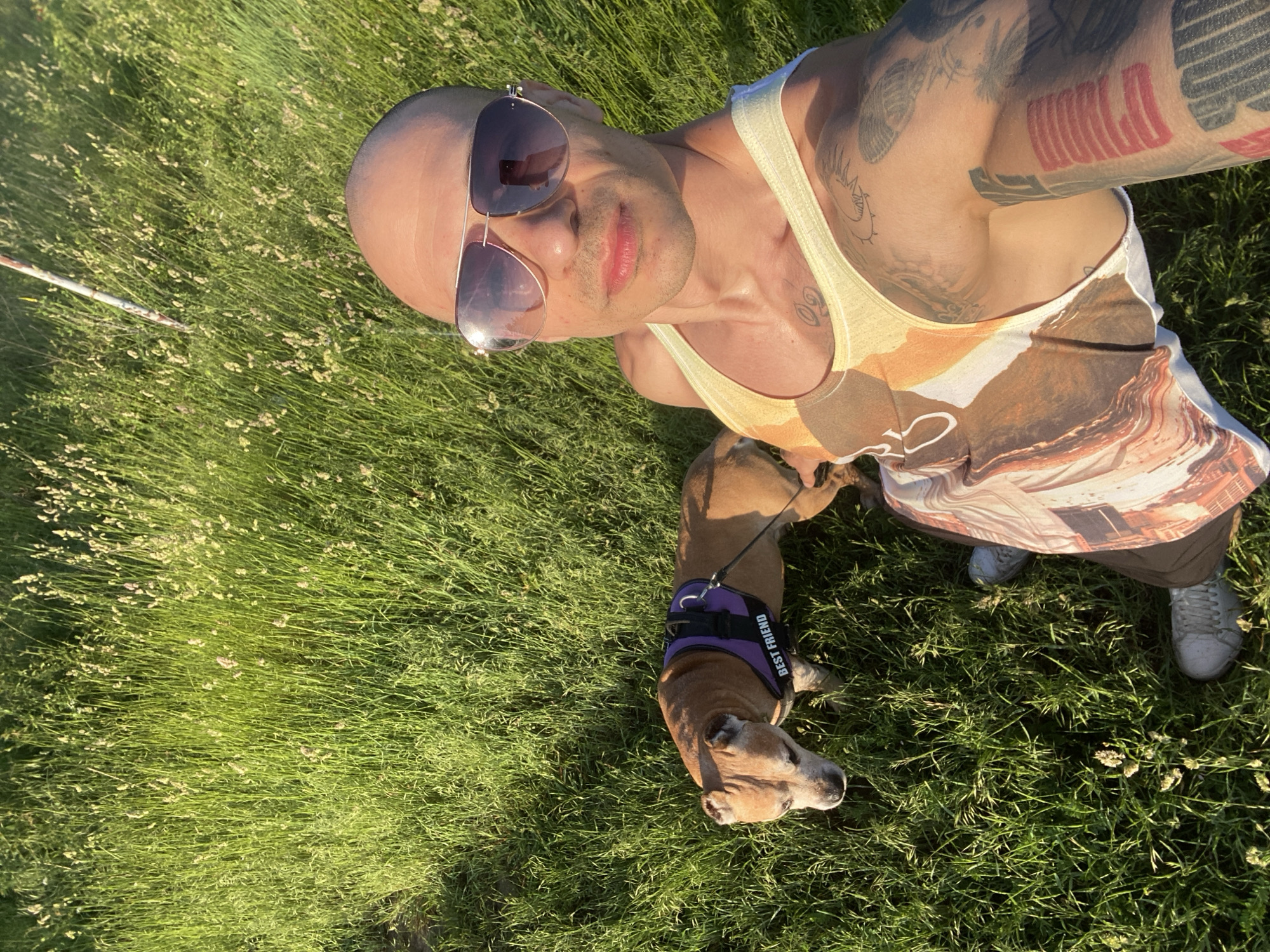 A person with tattoos, sunglasses, and a tank top is taking a selfie outdoors. The person is standing in tall green grass, and a brown dog wearing a harness labeled "BEST FRIEND" is standing beside them. The person is wearing white sneakers and brown trousers. The location is unknown, and there is no indication it is in Pleven, Bulgaria.