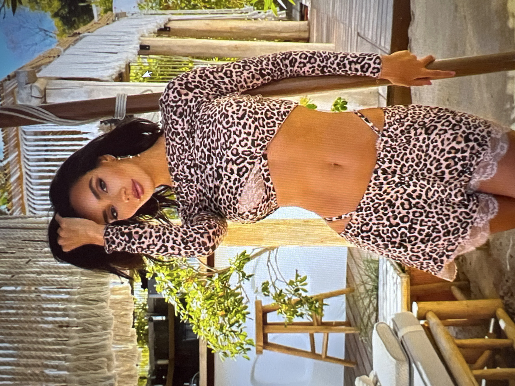 A woman stands on a wooden deck, her body angled towards the viewer. She wears a leopard-print two-piece outfit, consisting of a long-sleeved crop top and high-waisted shorts with lace trim. Her right hand is raised, holding a lock of her dark hair, while her left hand rests on a wooden pole. The background features a beach-like setting with sand, greenery, and structures made of wood and rope. There is no visible indication that this location is in Sofia, Bulgaria.