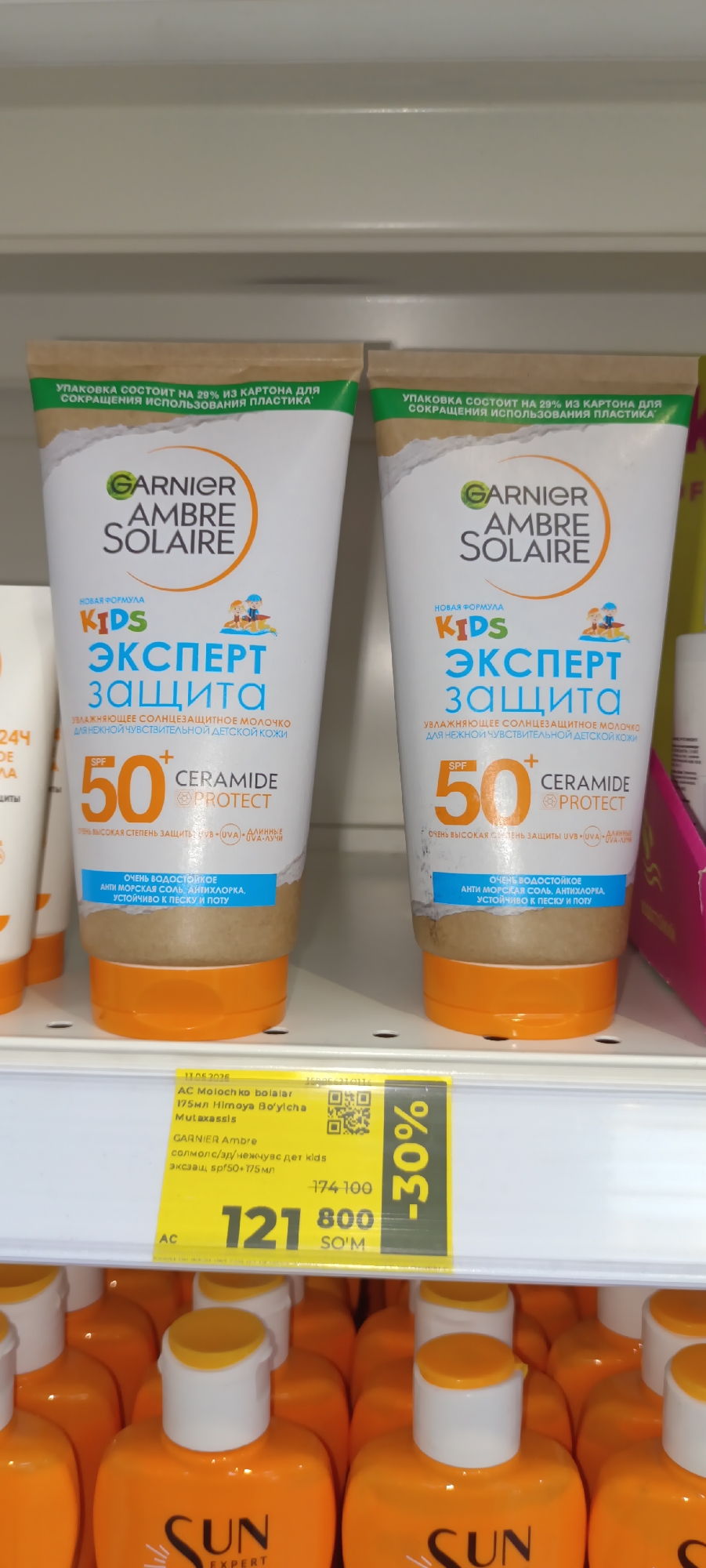 The image displays two tubes of Garnier Ambre Solaire Kids Expert Protection sunscreen with SPF 50, positioned side-by-side on a shelf in what appears to be a retail environment. The tubes are primarily white with orange caps and branding. Text on the tubes indicates "Kids Expert Protection," "UVA+UVB protection," and "Ceramide Protect." Below the tubes, a yellow price tag is visible with text in a foreign language, indicating a sale of "30%". The price tag shows "174 100" crossed out and "121 800" with "SO'M" underneath. Also on the price tag, it reads "AC Molochko bolalar", "175 ml Himoya Bo'yicha Mutaxassis", and "GARNIER Ambre solmolc/3p/неживусдет kids экczaш sp'50+175 мл". The overall scene suggests a product display for sunscreen on a store shelf. The image does not provide any specific information about the city of Fergana, Uzbekistan, or any individuals present.
