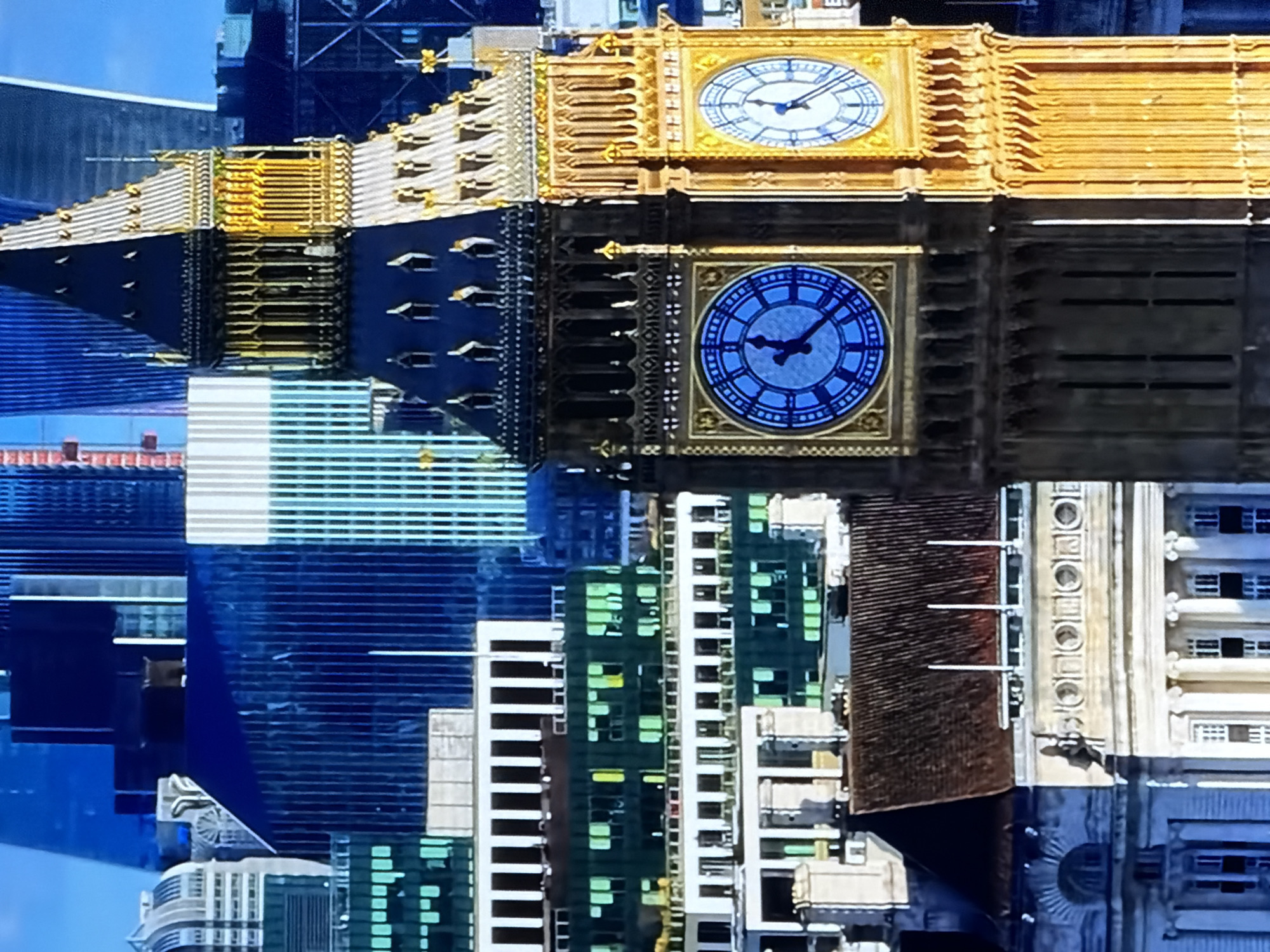 The image captures a detailed, telephoto view of the Elizabeth Tower, commonly known as Big Ben, situated in London, United Kingdom, not Maidstone. The tower occupies the right side of the frame, showcasing its golden upper sections and two prominent clock faces. The clock face on the left side of the tower displays a time of approximately 11:55, with its numerals and hands illuminated in a striking blue against a darker blue background. The clock face on the right, bathed in warm, golden sunlight, shows a time closer to 11:50, with its details in lighter blue against a white background.
Behind and to the left of the tower, a dense urban landscape unfolds under a clear blue sky. Numerous modern skyscrapers with reflective glass facades dominate the background, appearing in shades of blue and grey. Some mid-rise buildings are visible with prominent green-tinted windows. In the lower-left portion of the frame, older, lighter-colored architectural elements with traditional windows and a brown tiled roof are partially visible, contrasting with the contemporary structures. The lighting suggests bright daylight, with strong shadows and a golden illumination on the right side of the Elizabeth Tower. No people are discernible in the scene.