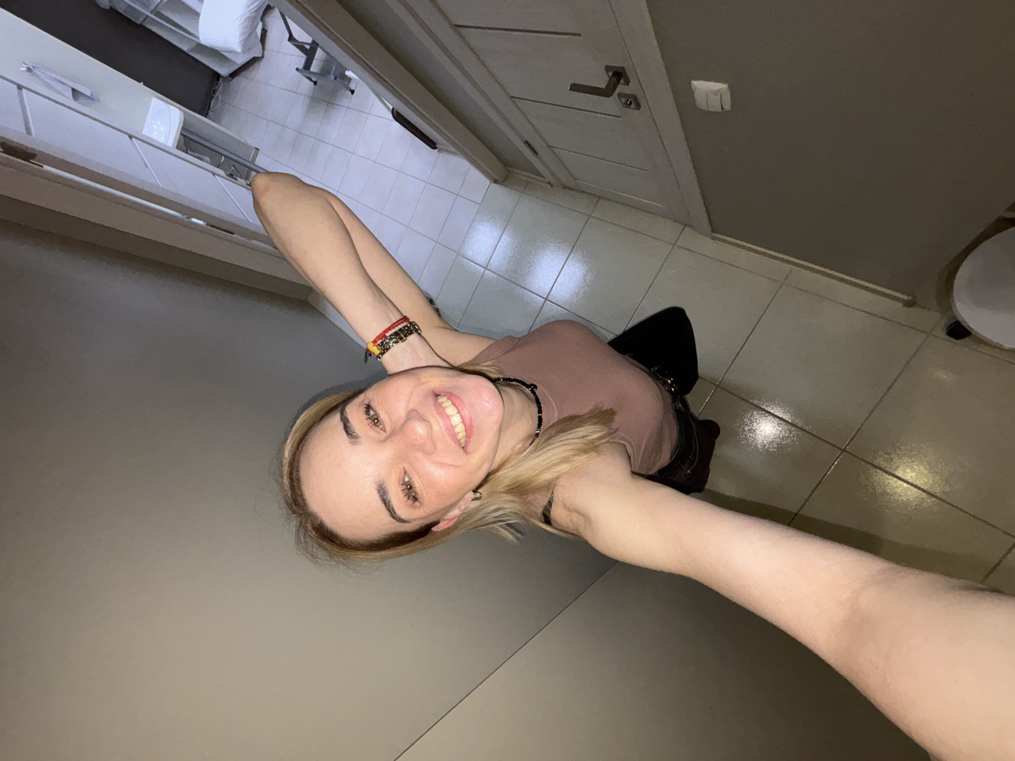 The image is a selfie taken from a low angle. A young woman with fair skin and blonde hair is smiling at the camera. She is wearing a brown tank top, black pants, and bracelets on her left wrist. She also has a black necklace. The background includes a tiled floor, a white door with a silver handle, a light switch, and a gray wall. In the distance, there is a glimpse of what appears to be a hospital bed or examination table. The location is identified as Unknown, Russia.