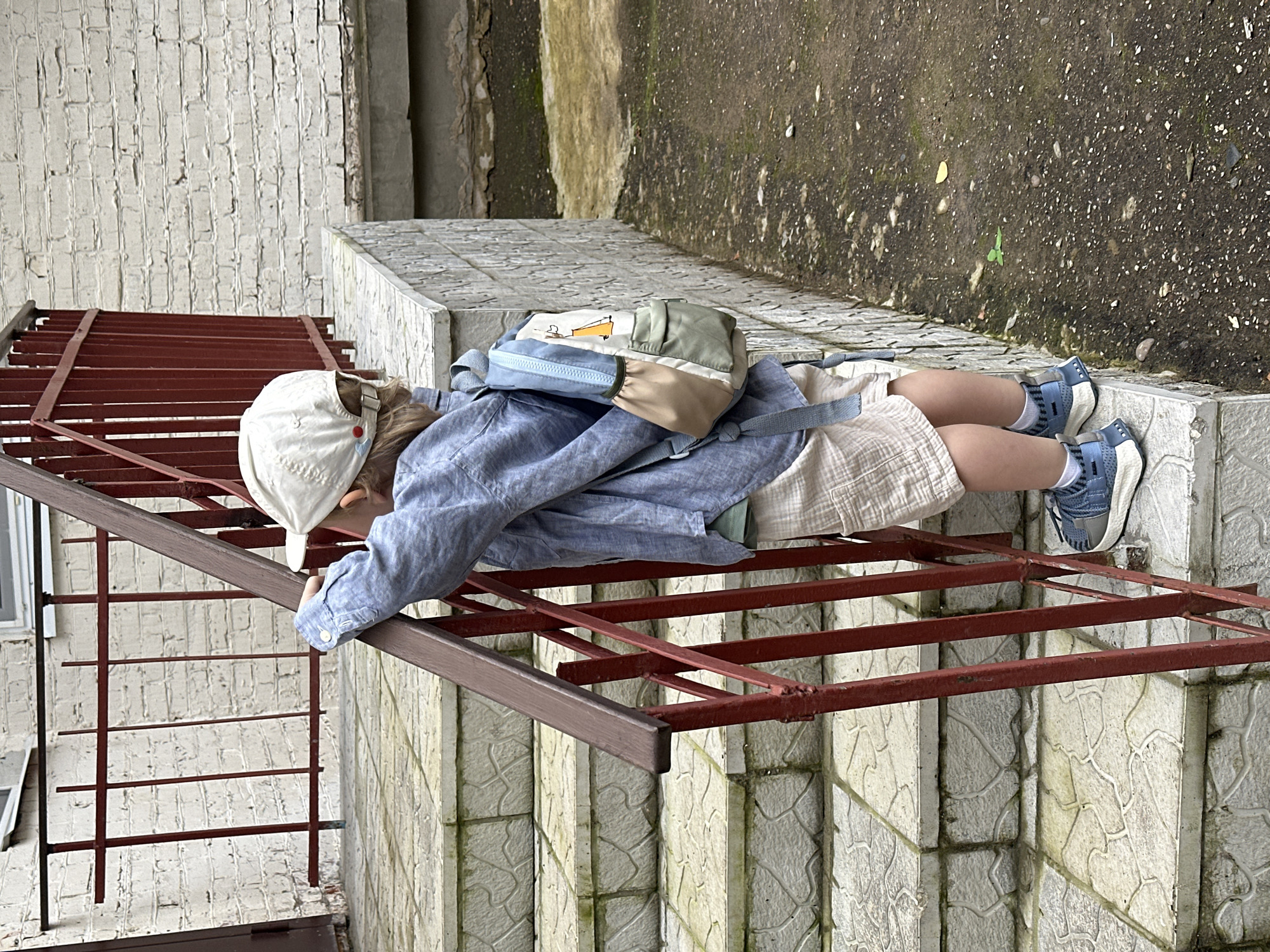 A person, likely a child, is lying prone on a rusty metal structure, possibly a fire escape or scaffolding, with their head and upper body on the structure and their legs extending downwards. They are wearing a blue collared shirt, light-colored shorts, white socks, and blue athletic shoes. A light-colored baseball cap covers their head, and a backpack with multiple pockets in green, tan, and light blue is worn on their back. The person appears to be resting or observing their surroundings.
The scene is outdoors, with a textured concrete wall and a rough, pebbled ground visible in the background. The wall has patches of moss and dirt, suggesting it's an older or weathered structure. The ground also appears damp in places. The metal structure is made of square tubing and has a reddish-brown hue, showing signs of rust and age. The overall impression is an urban or semi-urban environment, possibly a backyard, alley, or industrial area. The lighting suggests it might be daytime, but the specific time is unclear due to the overcast or diffused light. There is no visible text in the image.