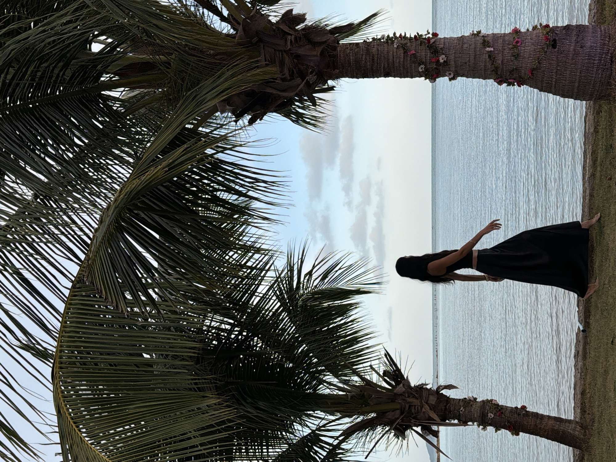In Ko Samui District, Thailand, a solitary woman with long, dark hair walks along a grassy shoreline beside the ocean. She wears a black sleeveless top and a long black skirt. Her body is positioned in profile, facing away from the viewer and towards the water. Two palm trees frame the scene, with their fronds reaching towards a cloudy sky. The palm tree on the right is decorated with a vine of pink and white flowers. The ocean stretches out to the horizon, and a few indistinct structures are visible in the distance. The overall atmosphere is serene and natural.