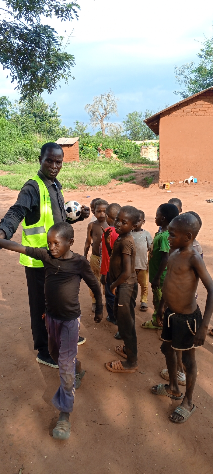 The image portrays an outdoor scene in Ngalula, Democratic Republic of the Congo, featuring one adult male and a group of approximately nine to ten children on a dry, reddish-brown dirt surface. The adult male, positioned on the left side of the frame, wears a dark long-sleeved shirt with a neon yellow reflective vest over it, dark trousers, and dark footwear. He holds a black and white soccer ball in his left hand and extends his right arm to the left, gesturing towards a child. The children, all of African descent and appearing to be between five and ten years old, are clustered in the center and right. Their clothing varies; some are shirtless, while others wear t-shirts in colors such as red, green, brown, and grey, paired with shorts or trousers. Footwear includes sandals, simple shoes, or bare feet. In the background, two structures constructed from reddish-brown mud bricks are visible, with dense green vegetation and several trees, including a prominent light-colored, possibly dormant tree, under a light blue sky with scattered clouds.