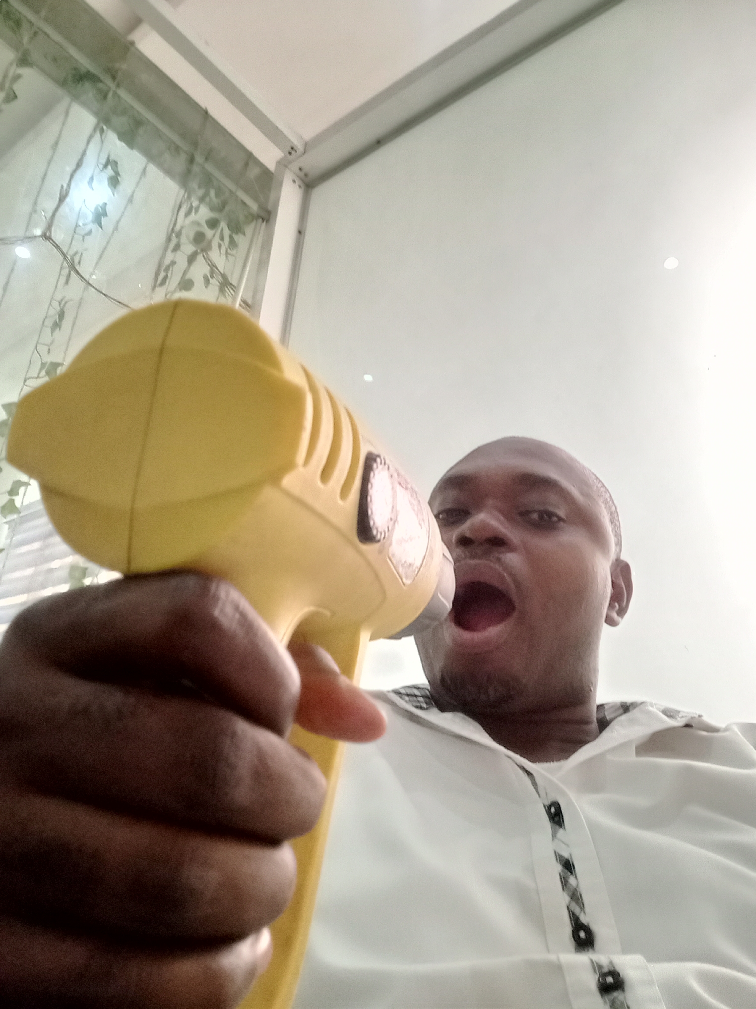 A man is holding a yellow toy megaphone toward the camera, with his mouth open as if he is shouting. The man is dark-skinned, with short hair and is wearing a white shirt. The background shows a room with a window covered by a light green curtain with a leaf pattern. The location is Gwarinpa, Nigeria.