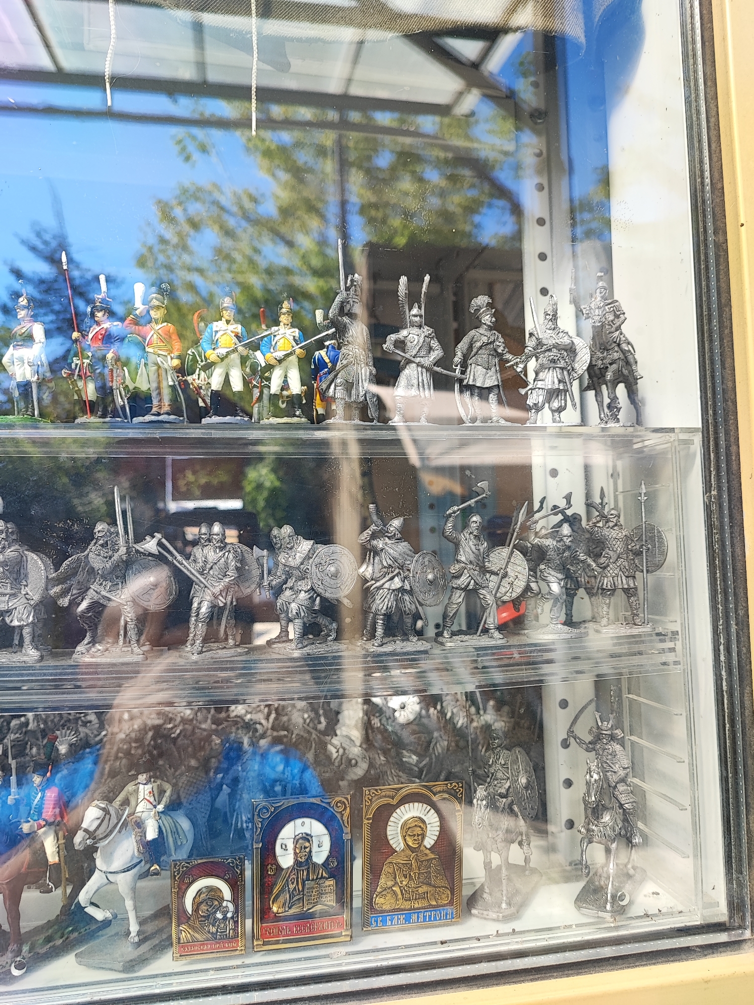 This image shows a display case with miniature figurines of soldiers and religious icons. The display case is made of clear plastic and is set against a backdrop of trees and a blue sky, indicating it is outdoors or near a window. The figurines are arranged on multiple shelves. The top shelf features brightly colored figurines of soldiers in historical uniforms, holding rifles and spears. The middle shelf contains silver-colored figurines of warriors in medieval armor, some with shields and swords. The bottom shelf displays more silver-colored figurines of warriors, including one on horseback in samurai armor, and several religious icons with gold and enamel detailing. The overall scene suggests a shop or a collector's display. The image was taken in Pyatigorsk, Russia.