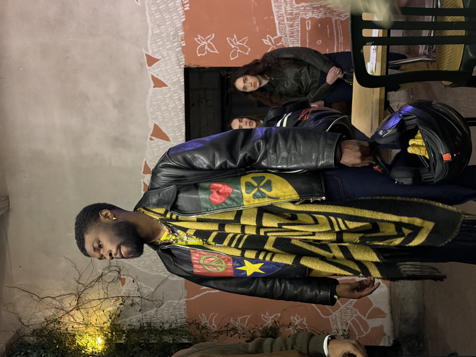An adult black male stands prominently in the foreground of an indoor or semi-outdoor setting in Bagnolet, France. He has short, textured dark hair, a full beard and mustache, and a visible gold hoop earring in his left ear. He wears a black leather jacket adorned with four square patches: a red patch with a green peace sign (top left), a light green patch with a red heart (top right), a dark blue patch with a yellow star (bottom left), and a yellow patch with a green four-leaf clover (bottom right). A thick yellow and black striped scarf is draped around his neck, concealing a yellow undershirt. Multiple necklaces, including one with a silver-colored crucifix pendant, are visible. He wears dark blue trousers and black beaded bracelets on his left wrist. In his right hand, he holds a black motorcycle helmet with bright yellow gloves visible inside. He is looking towards his left, with a neutral expression.
In the mid-ground, to the right of the main subject, two women are partially visible. One woman, with long dark hair and wearing a dark green jacket, stands with her hands clasped in front of her, looking forward. Another woman, with long dark hair and a black and white top, is partially obscured between the main subject and the first woman, her face turned slightly away.
The background consists of textured walls. The upper wall section is light gray/off-white, with a rough, torn border revealing a reddish-orange section below. This lower reddish-orange wall features white outline drawings, resembling flowers or leaves. A string of reddish-orange triangular pennants or paper cutouts is strung along the border between the two wall sections. To the far left, green vines with small leaves grow up the wall, illuminated by a warm yellow light source. A light-colored wooden table with an object on it and a chair with a woven cane seat are visible to the right of the main subject. The floor is not distinctly visible. The lighting suggests an indoor or evening setting.
