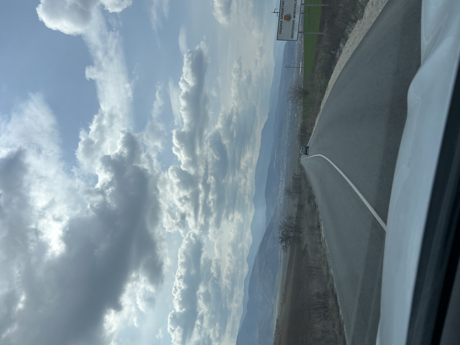 The image presents a forward-facing view from a white vehicle on a paved two-lane road, marked by a continuous white center line. The road extends towards a distant mountainous horizon under a partly cloudy sky. A silver vehicle proceeds ahead in the same direction on the road. On the right side of the road, a white rectangular sign displays "ОБЩИНА БРАЦИГОВО" and "BRATSIGOVO MUNICIPALITY", identifying the location as Bratsigovo Municipality, Bulgaria. The immediate roadside on both sides consists of dry, sparse vegetation and brown earth. A section of green cultivated field is visible further along on the right, beyond the sign. A small town is situated in the mid-distance valley, nestled between the foreground and the distant mountains. The sky above is partially covered by dispersed white and grey cumulus clouds against a blue background.