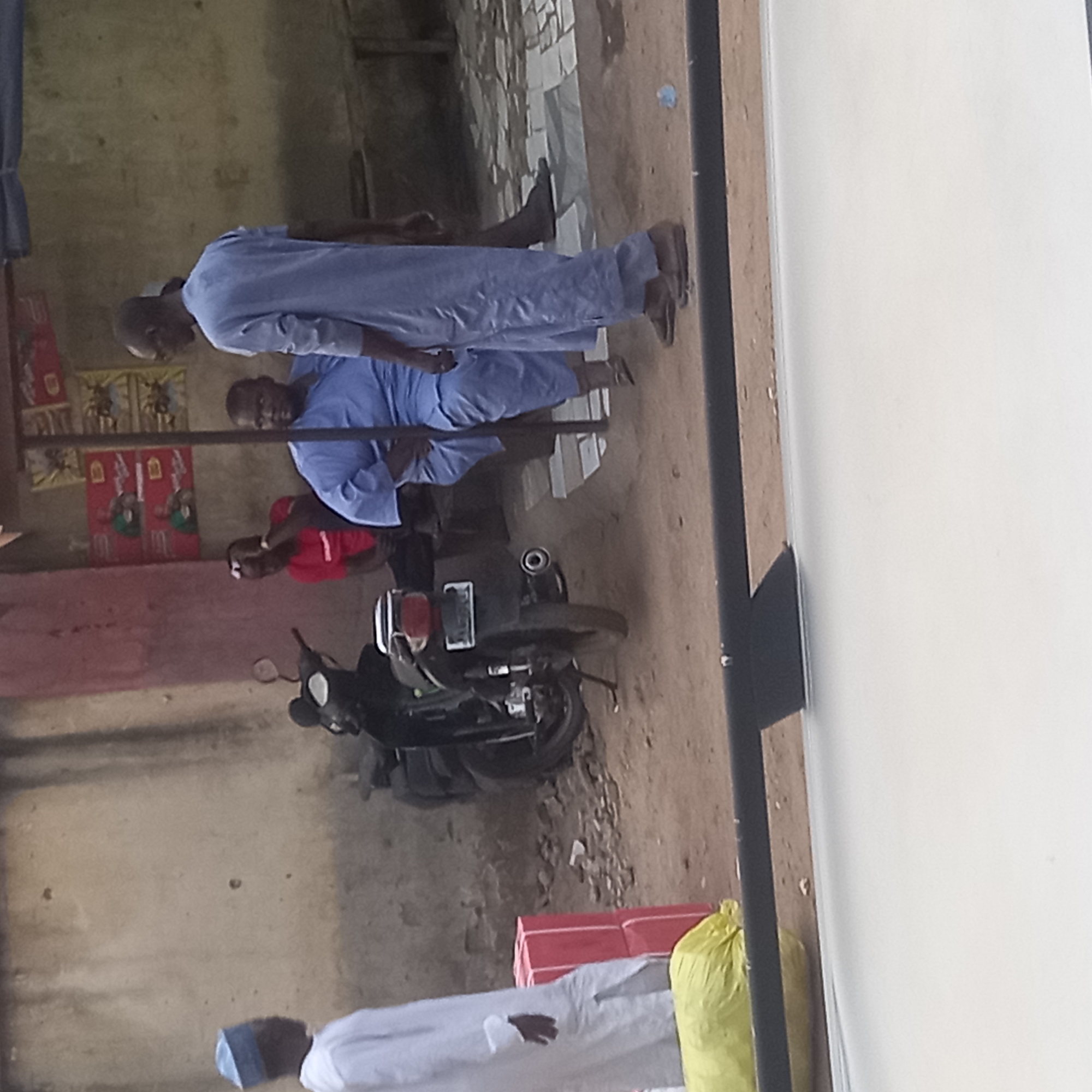 The scene depicts an outdoor gathering in Dadi Riba, Nigeria. Two men wearing light blue robes and caps stand near a motorcycle parked on a dirt ground. A man in a red shirt is seated next to the motorcycle. A man wearing a white garment and a blue cap is present near red crates and a yellow bag. The backdrop consists of a weathered building facade with faded posters.