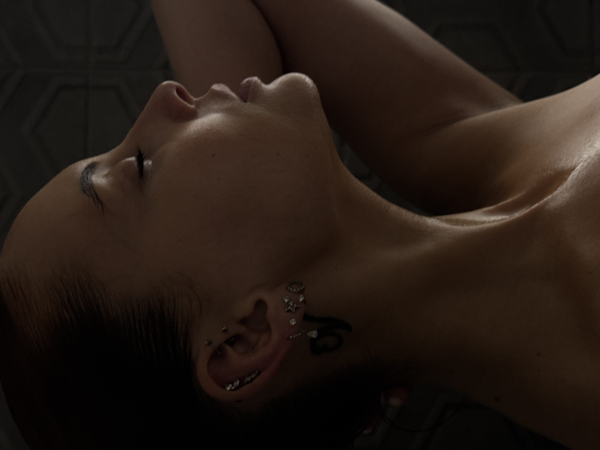 An intimate, low-light portrait captures a person in profile, likely female, reclining with their head tilted upwards and eyes closed or heavily lidded. The composition focuses on the subject's face, neck, and shoulder, which are softly illuminated from above, highlighting the smooth contours of the skin. The person's bare upper body suggests a state of rest or relaxation.
On the visible right ear, there are numerous piercings, including various studs and rings with designs such as stars and circles. A dark, stylized tattoo extends from behind the ear down onto the upper neck, appearing to be a symbolic design rather than legible text. Dark hair is pulled back, revealing the ear and neckline.
The background is dark and features a subtle geometric hexagonal pattern, possibly a wall or headboard, adding texture without distracting from the subject. The soft, directional lighting suggests a nighttime or dimly lit indoor setting, creating a serene and contemplative mood. The person's relaxed posture and closed eyes evoke a sense of peace or deep thought. No visible text is present in the image. While the provided location context is Chaikovsky, Russia, there are no specific visual cues within the image itself to confirm this location.