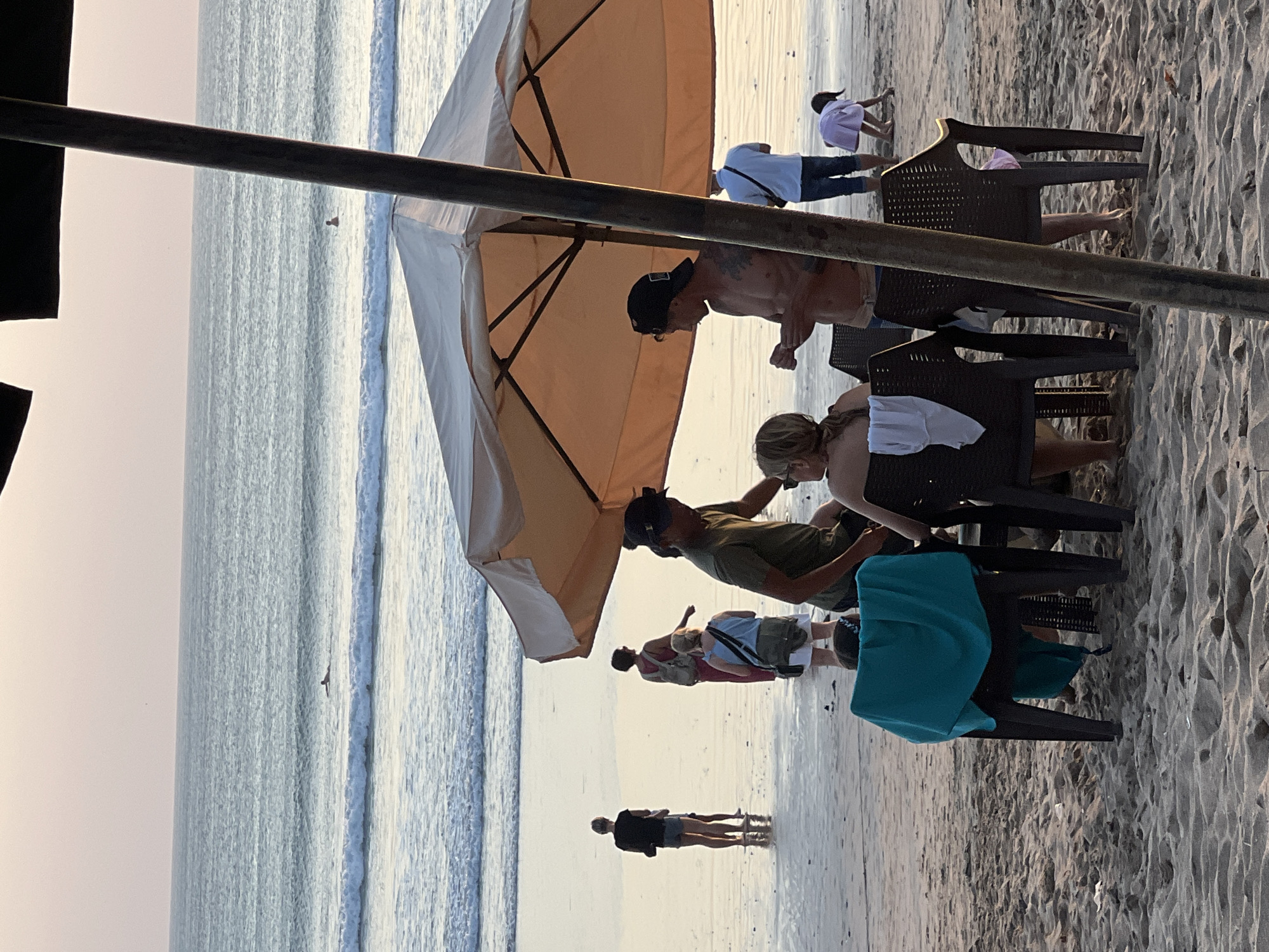 This image captures a serene sunset scene on Kuta Beach, Bali, Indonesia. The warm, golden light illuminates the wet sand where several individuals are present. In the foreground, two chairs are set up under a large, beige beach umbrella, with a turquoise towel draped over one. Nearby, a couple is engaged in conversation, the man shirtless and wearing a baseball cap, while the woman has her back to the camera.
Further down the beach, a group of people are walking near the water's edge. One woman in a pink dress is carrying a backpack, and another woman in a light blue top is walking with a bag. In the distance, a lone surfer glides across a gentle wave, silhouetted against the soft, pastel-colored sky. A small child, wearing a pink dress, is bent over, playing in the sand, while a man in a light blue shirt stands nearby. The ocean stretches to the horizon, with calm waves lapping the shore. The overall atmosphere is one of relaxation and quiet enjoyment of the beach at dusk.