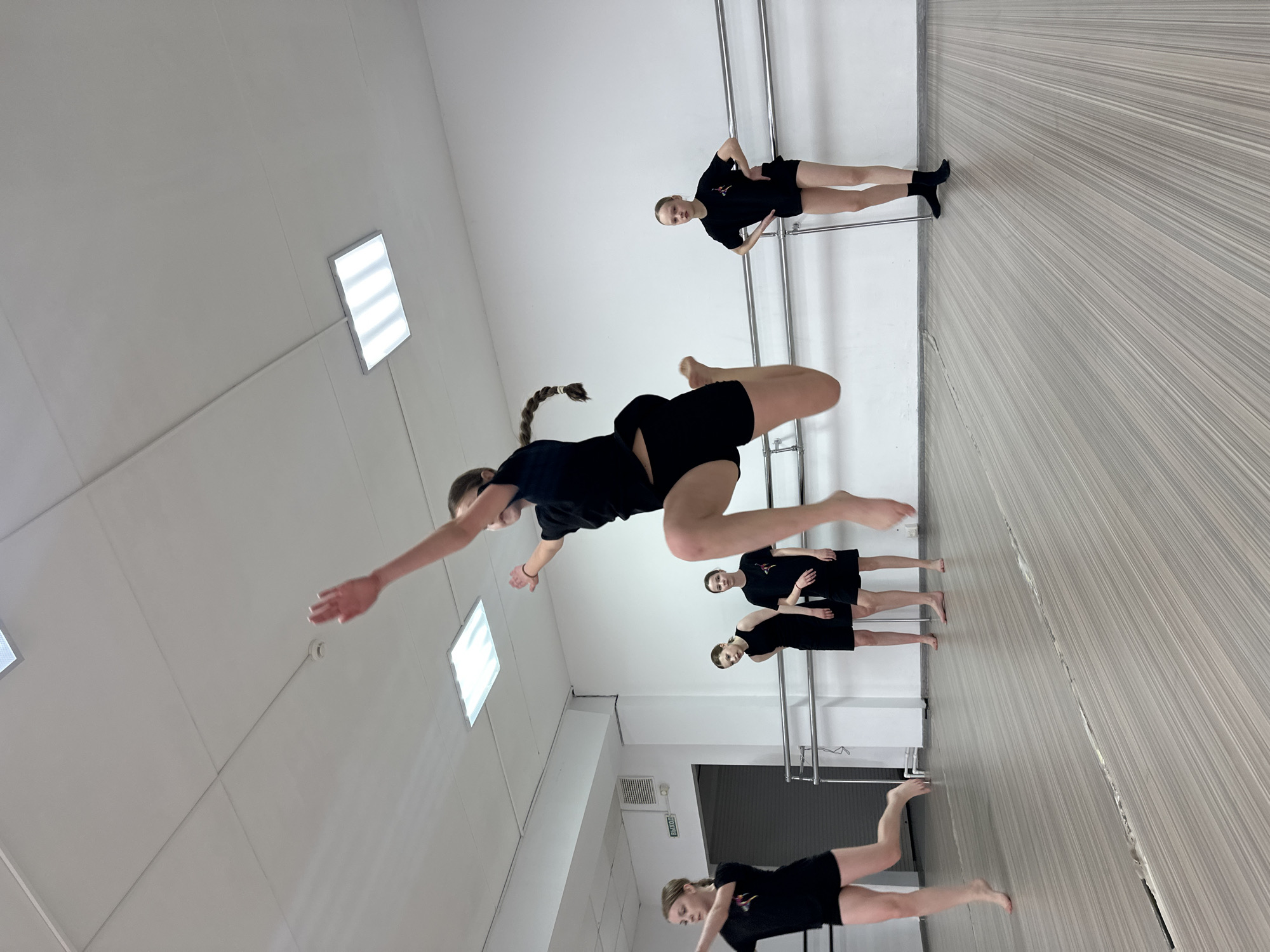 The image captures five young females engaged in a dance practice session within what appears to be a brightly lit studio. The overall scene is dynamic, dominated by a central figure in mid-air performing a powerful leap or jump. She is dressed in a black short-sleeved top and black shorts, with her arms extended and a braided ponytail trailing behind her.
In the background, three other dancers, similarly dressed in black tops and shorts, are standing near a silver-colored ballet barre. One of these dancers, positioned further back, is facing the viewer with hands on her hips. The other two are slightly to the right, observing or awaiting their turn. A fifth dancer is partially visible in the bottom right corner, lying or posing on the floor, suggesting another dance movement.
The setting is an indoor space with white walls and a light-colored wooden floor that exhibits a subtle striped pattern, also visible on a wall. The ceiling features rectangular fluorescent light panels, providing ample illumination. A small, round smoke detector or similar fixture is visible on the ceiling. A door with a sign reading "ВЫХОД" (VYKHOD), meaning "EXIT" in Russian, is visible in the background, confirming the location context of Votkinsk, Russia. The atmosphere suggests concentration and physical activity, typical of a dance rehearsal. No specific time of day or weather conditions are discernible from the indoor scene, but the bright artificial lighting suggests a well-lit environment.