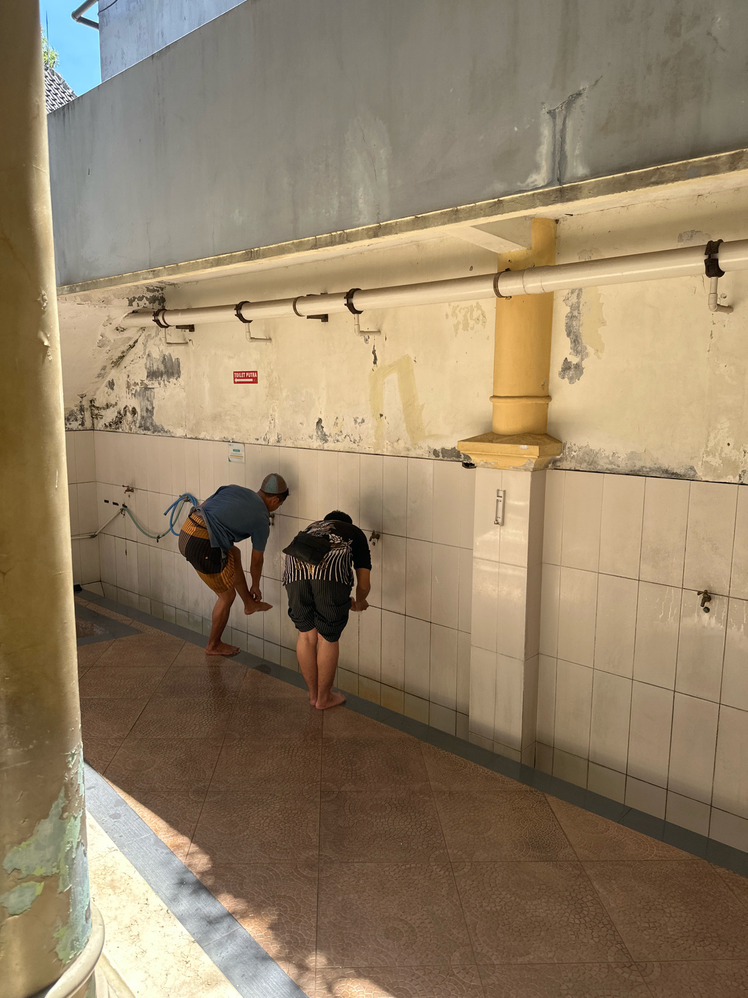 In Malang, Indonesia, two individuals are engaged in ritual ablutions in an outdoor washing area. The scene is characterized by tiled walls and floor, with a prominent yellow pipe extending vertically. The first individual, wearing a blue t-shirt and striped shorts, is facing the wall and appears to be washing their feet. The second individual, in a black t-shirt and striped shorts, is also facing the wall, presumably performing similar ablutions. Several water taps are visible along the wall, with one individual interacting with a tap. A red sign with white text indicating "TOILET PUTRA" (Male Toilet) with an arrow pointing left is affixed to the wall. The environment suggests a public restroom or a designated area for religious cleansing.