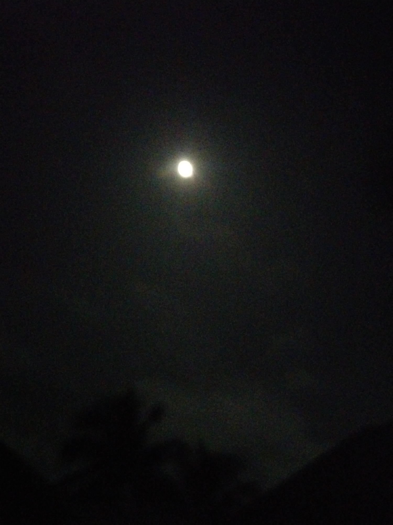 The image depicts a dark, nighttime scene dominated by a full moon against a black sky. The moon is a bright, circular object positioned slightly above the center of the frame. In the lower portion of the image, the dark silhouettes of trees and possibly hills are visible, suggesting an outdoor environment. The overall atmosphere is one of quiet darkness, with the moon being the sole significant light source. The image does not contain any elements that can be used to identify the specific location within Uyo, Nigeria.