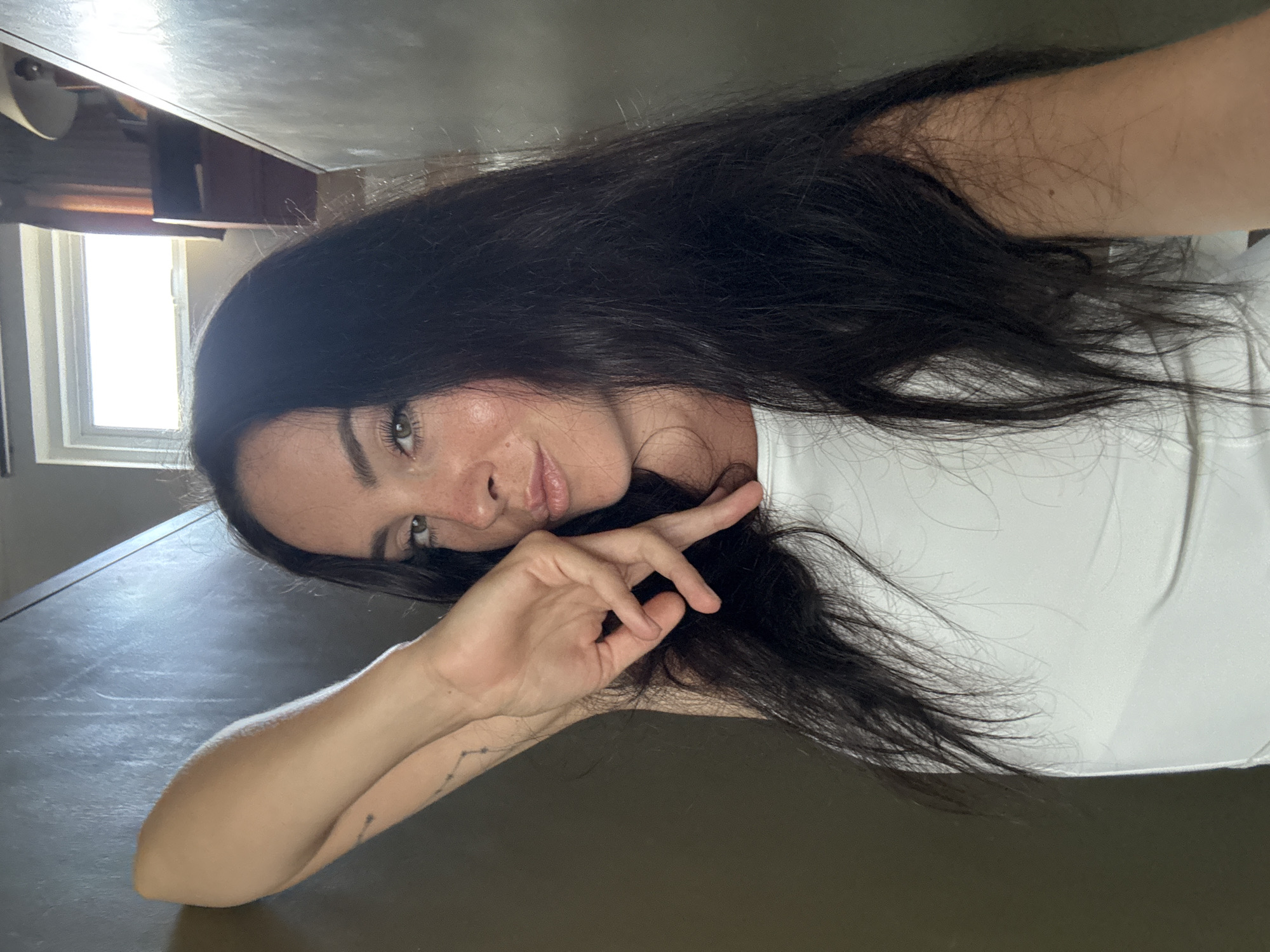 The image shows a woman with long, dark hair and light skin. She is wearing a white tank top. Her left arm is raised, with her hand positioned near her face. She is looking at the camera with a slight smile and is wearing makeup. There is a subtle tattoo on her left forearm. The background appears to be an indoor setting with neutral-colored walls, and a window with bright light visible in the upper right corner. There is no indication that this location is in North Kuta, Indonesia.