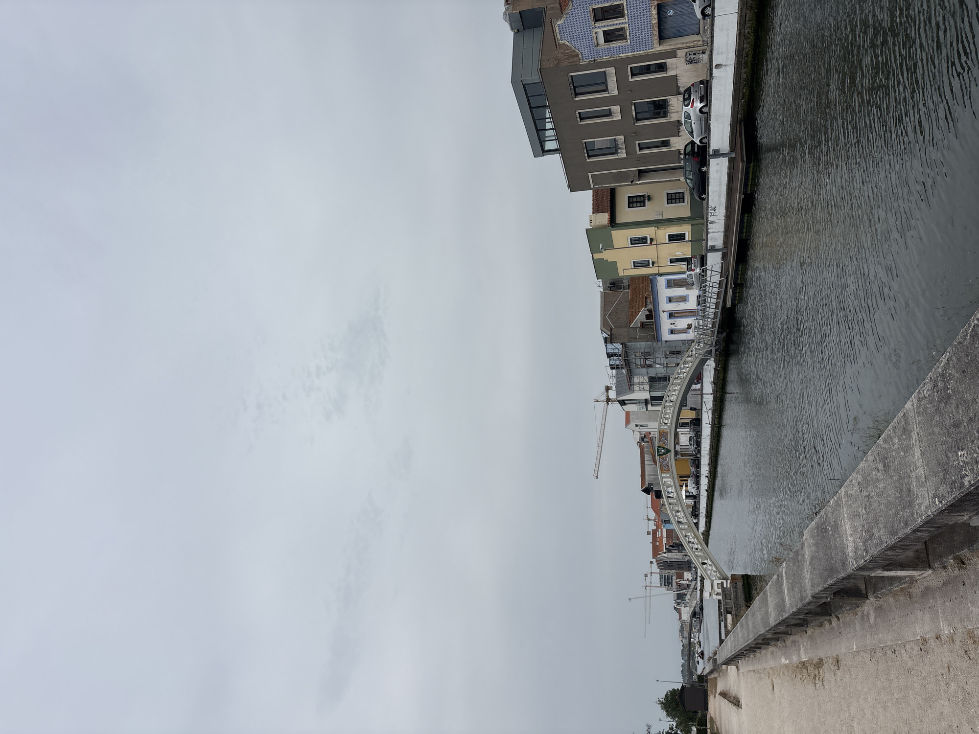 The image shows a canal in Aveiro, Portugal, with buildings lining the right bank and a paved walkway and stone wall on the left. The canal is calm and reflects the overcast sky. Several buildings of various architectural styles and colors are visible, including some with traditional blue and white tile facades. A white arched bridge spans the canal in the mid-ground, connecting the two sides. In the background, construction cranes indicate ongoing development. Cars are parked along the street behind the buildings on the right. The overall impression is a picturesque urban waterway scene under a cloudy sky.