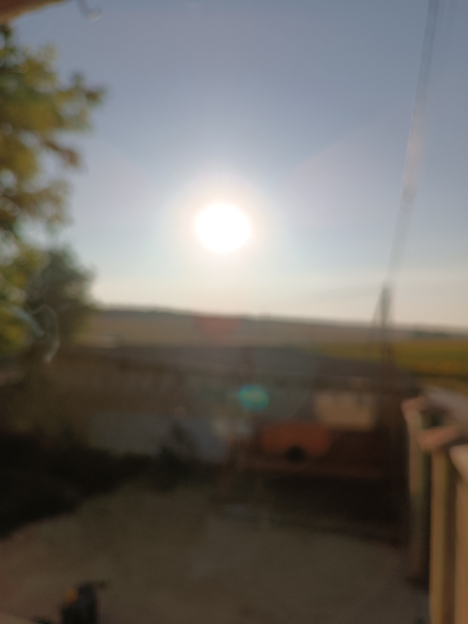 The image is a blurred, outdoor scene, likely taken in Copăceni, Moldova, during daylight. The sun is a bright, prominent orb in the upper center of the sky, creating lens flare. The sky is a pale blue, fading to a lighter hue near the horizon. In the middle ground, a blurred landscape of fields and structures is visible. A thin, dark, vertical line, possibly a pole or wire, is present in the upper right quadrant of the image. The foreground is dark and indistinct, with a suggestion of a railing or fence on the right side, and a blurred tree on the left. The overall impression is of a hazy, sunlit outdoor environment.